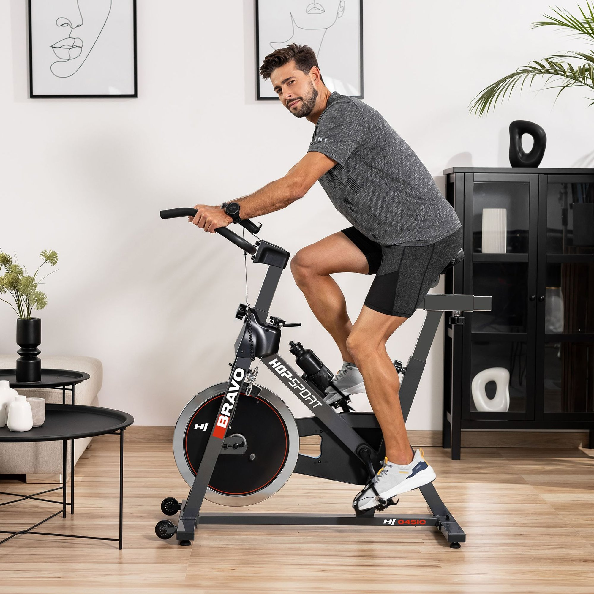 Ein Mann in Sportkleidung sitzt auf einem schwarzen Heimtrainer in einem modernen Wohnzimmer.