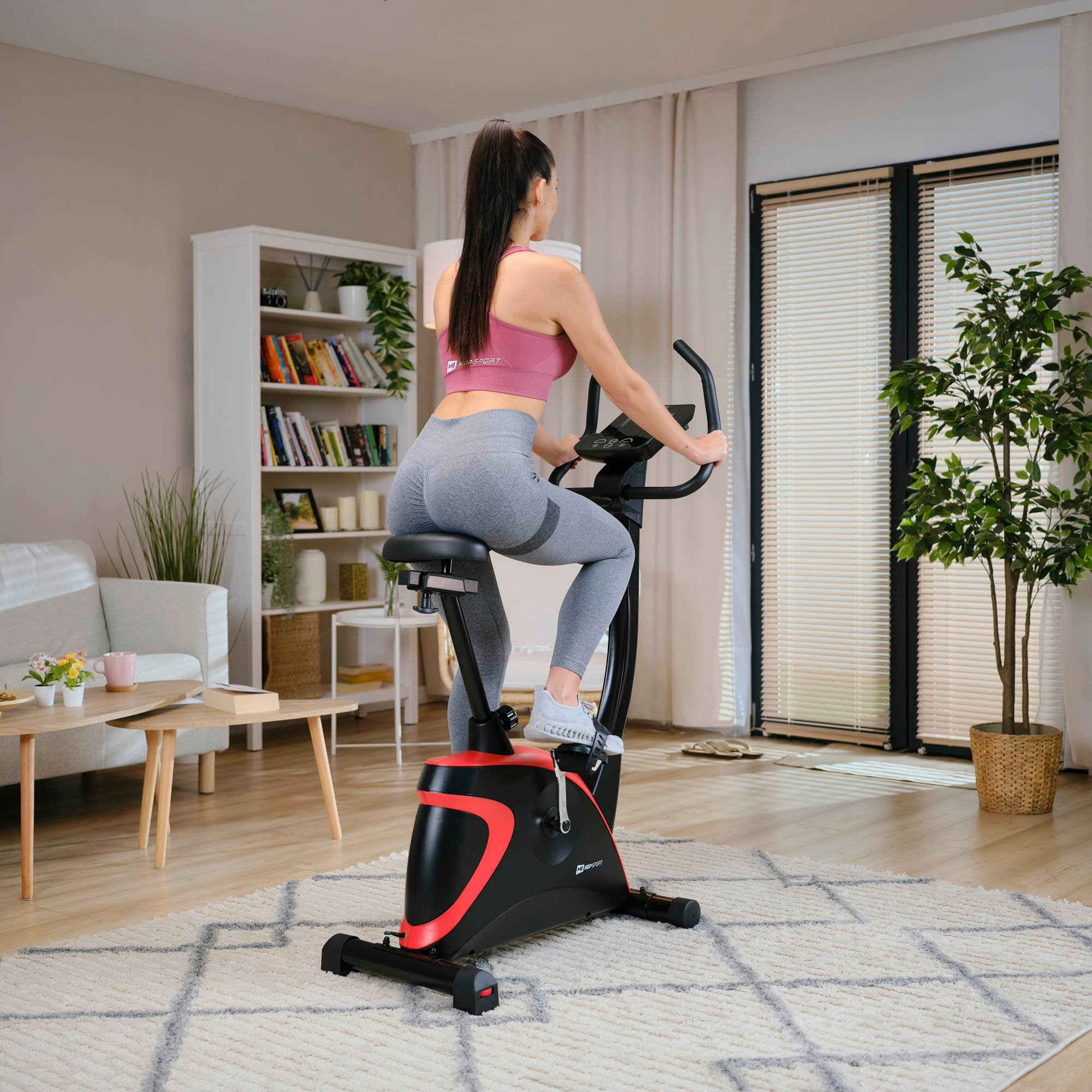 Eine Frau mit pinkem Sport-Top und grauen Leggings trainiert zu Hause auf einem schwarzen Heimtrainer.