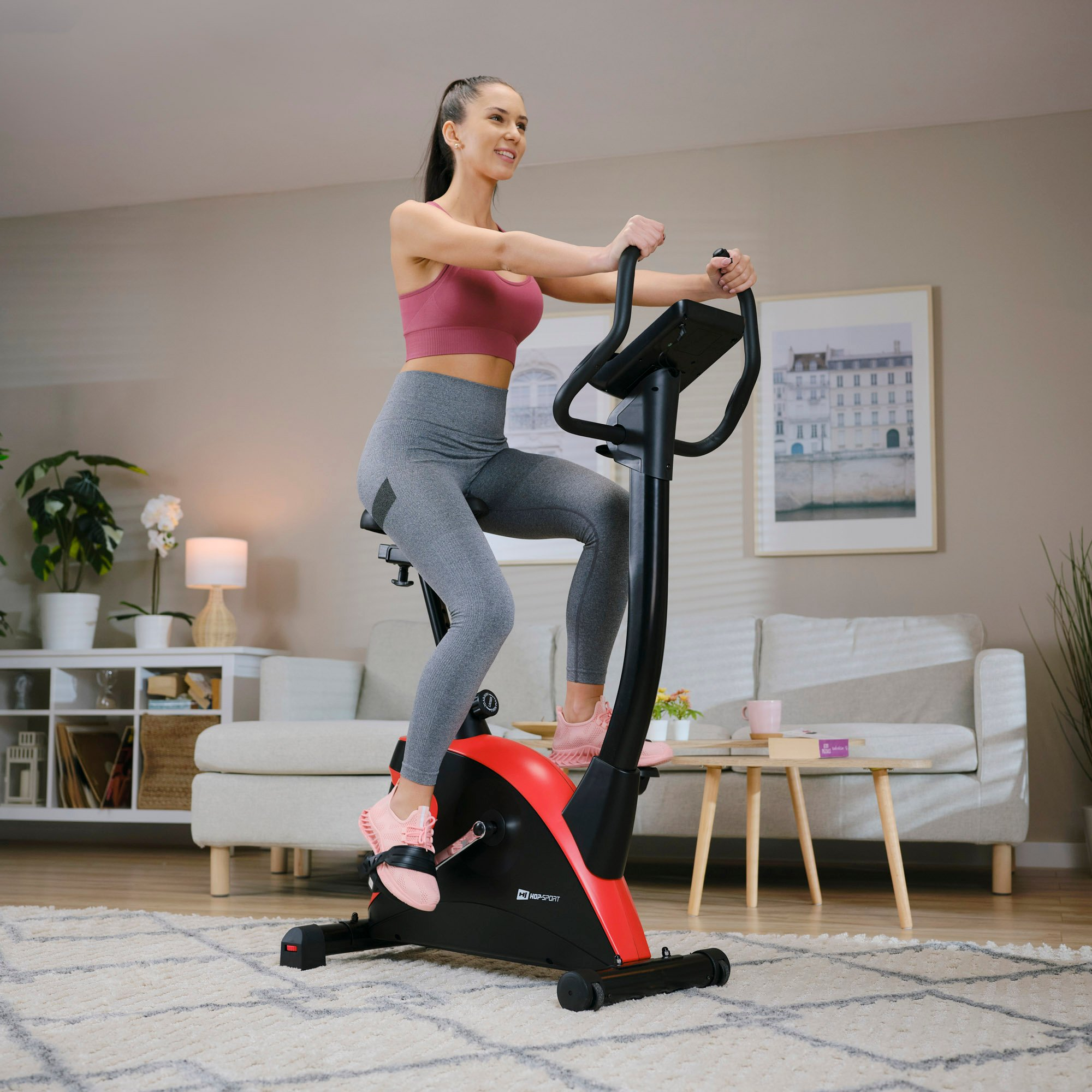 Eine Frau in Sportkleidung trainiert auf einem schwarzen und roten Heimtrainer im Wohnzimmer.
