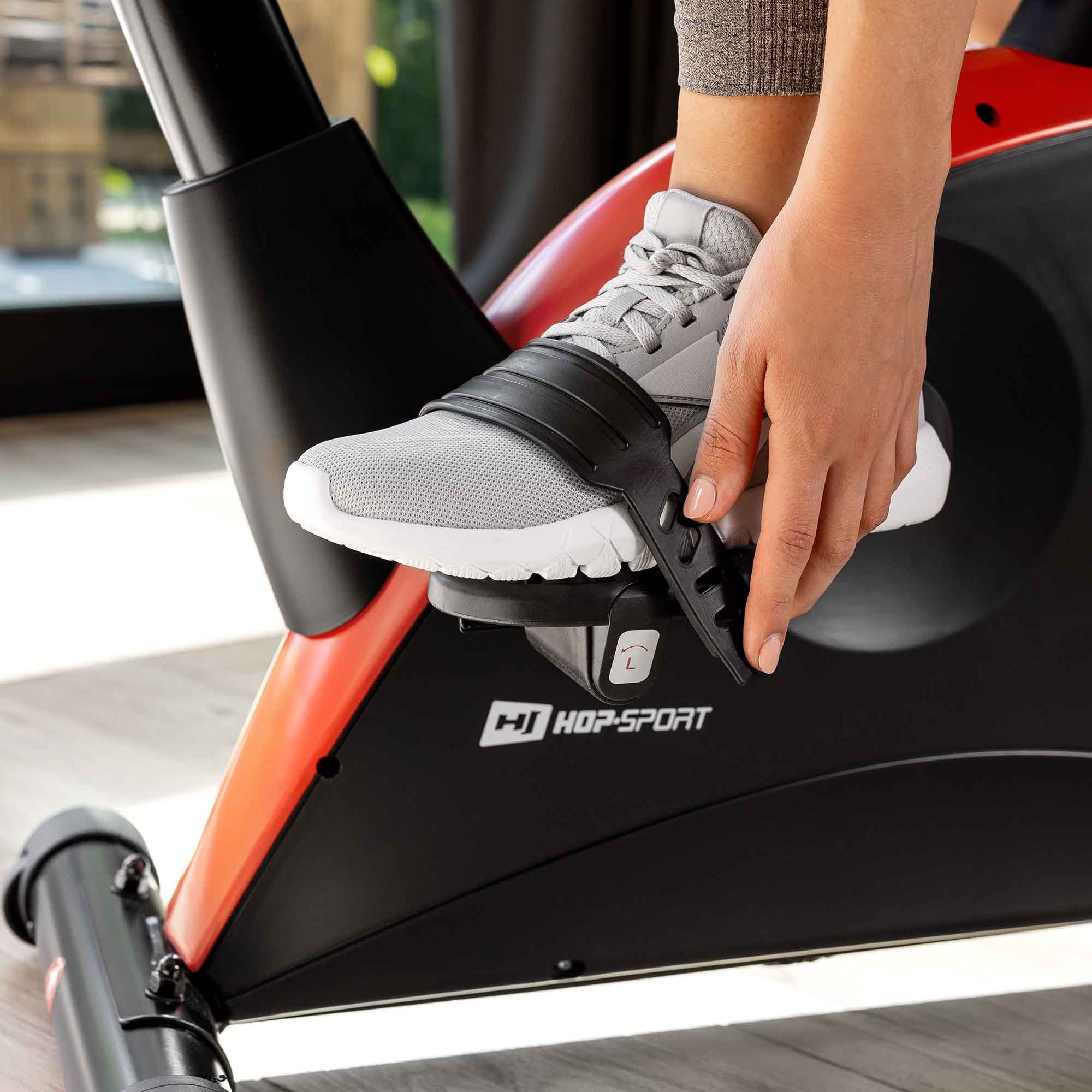 Eine Hand sichert einen grauen Sportschuh mit Riemen auf dem Pedal eines Heimtrainers.