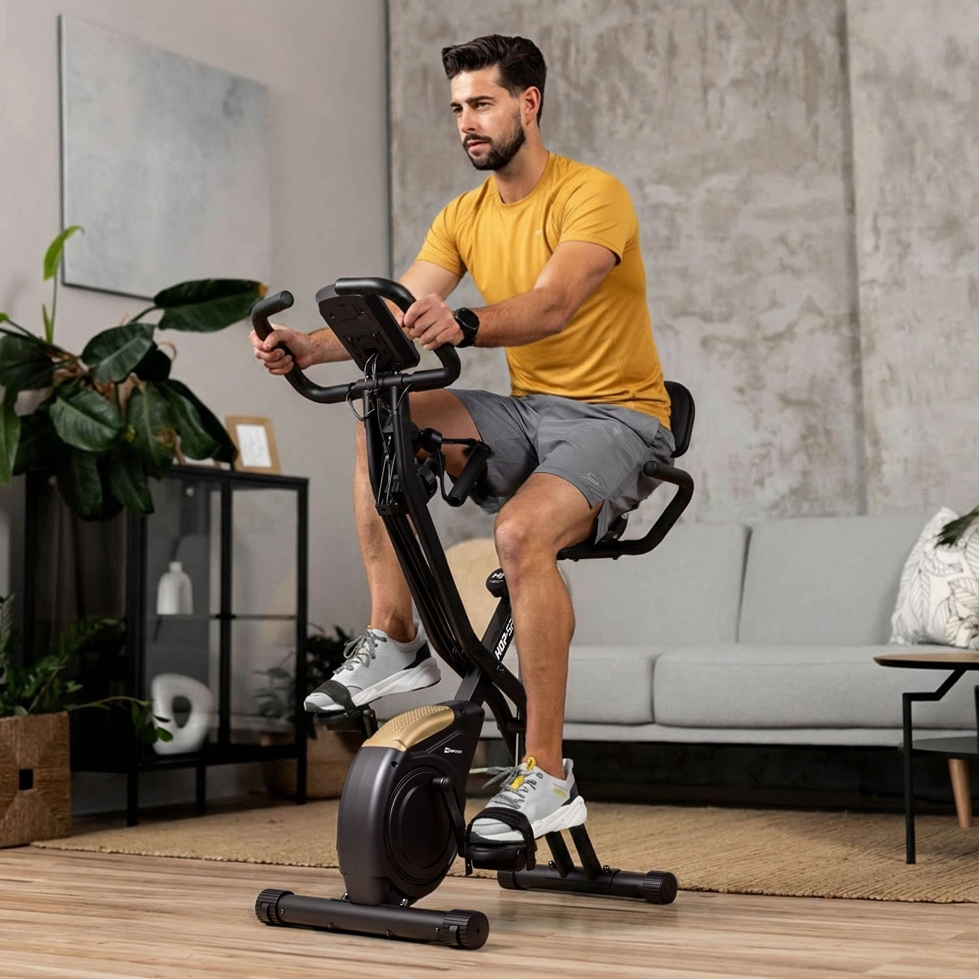 Ein Mann im gelben T-Shirt trainiert auf einem Heimtrainer in einem modernen Wohnzimmer.