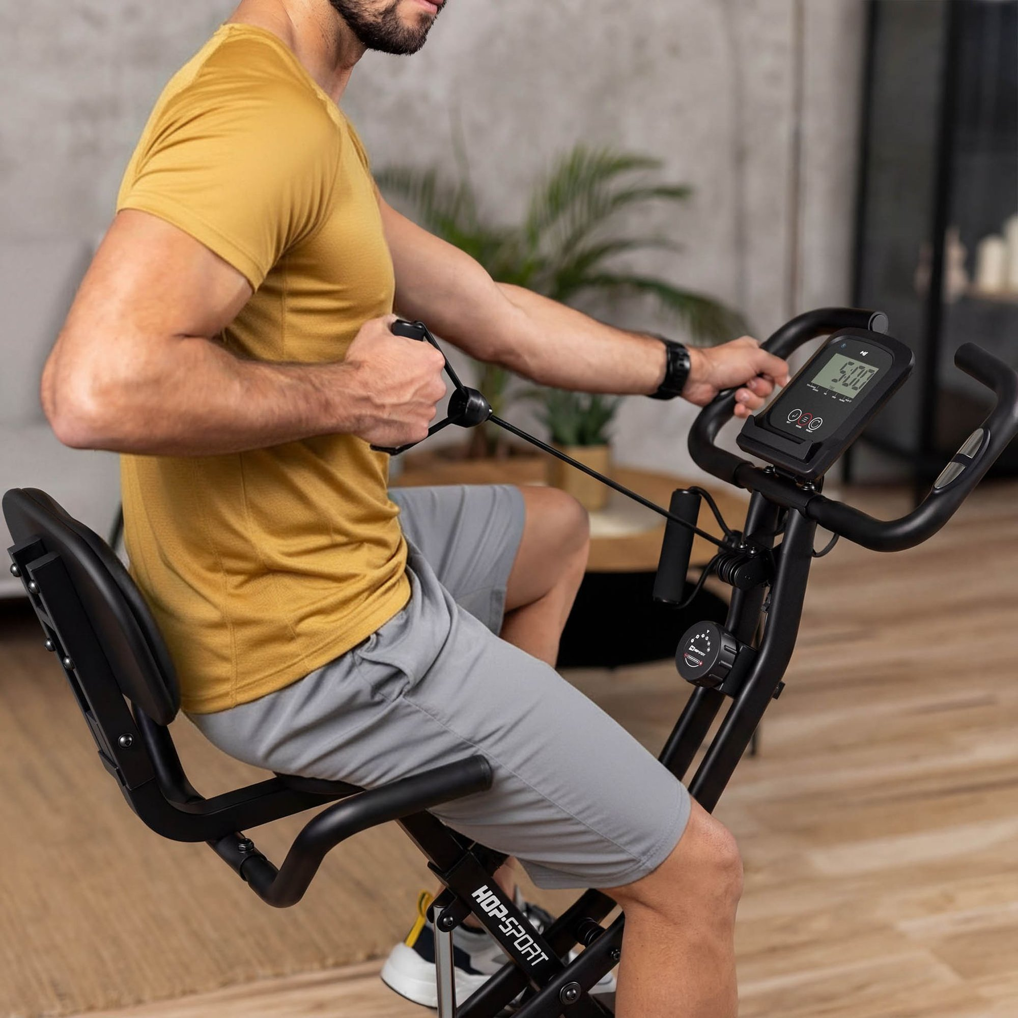 Ein Mann im gelben T-Shirt trainiert auf einem Heimtrainer mit Widerstandsbändern in einem Raum.