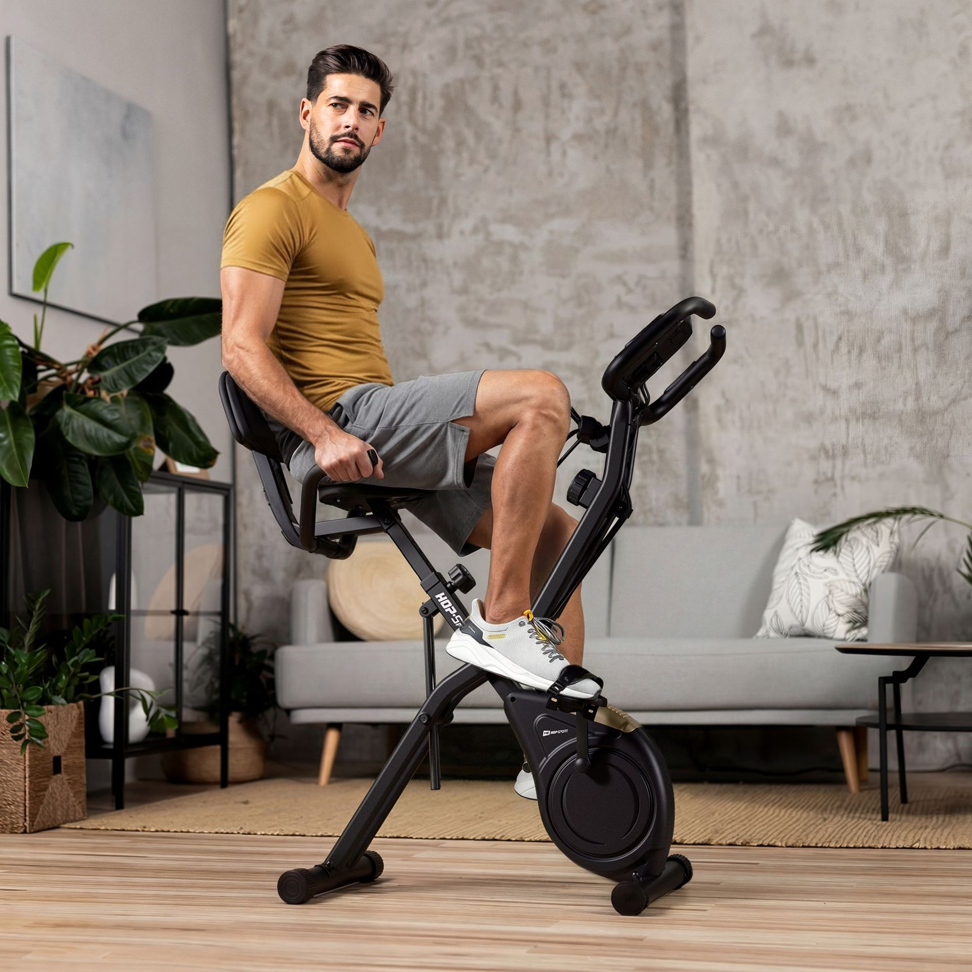 Ein Mann sitzt in einem modernen Wohnzimmer auf einem schwarzen Heimtrainer und blickt zur Seite.