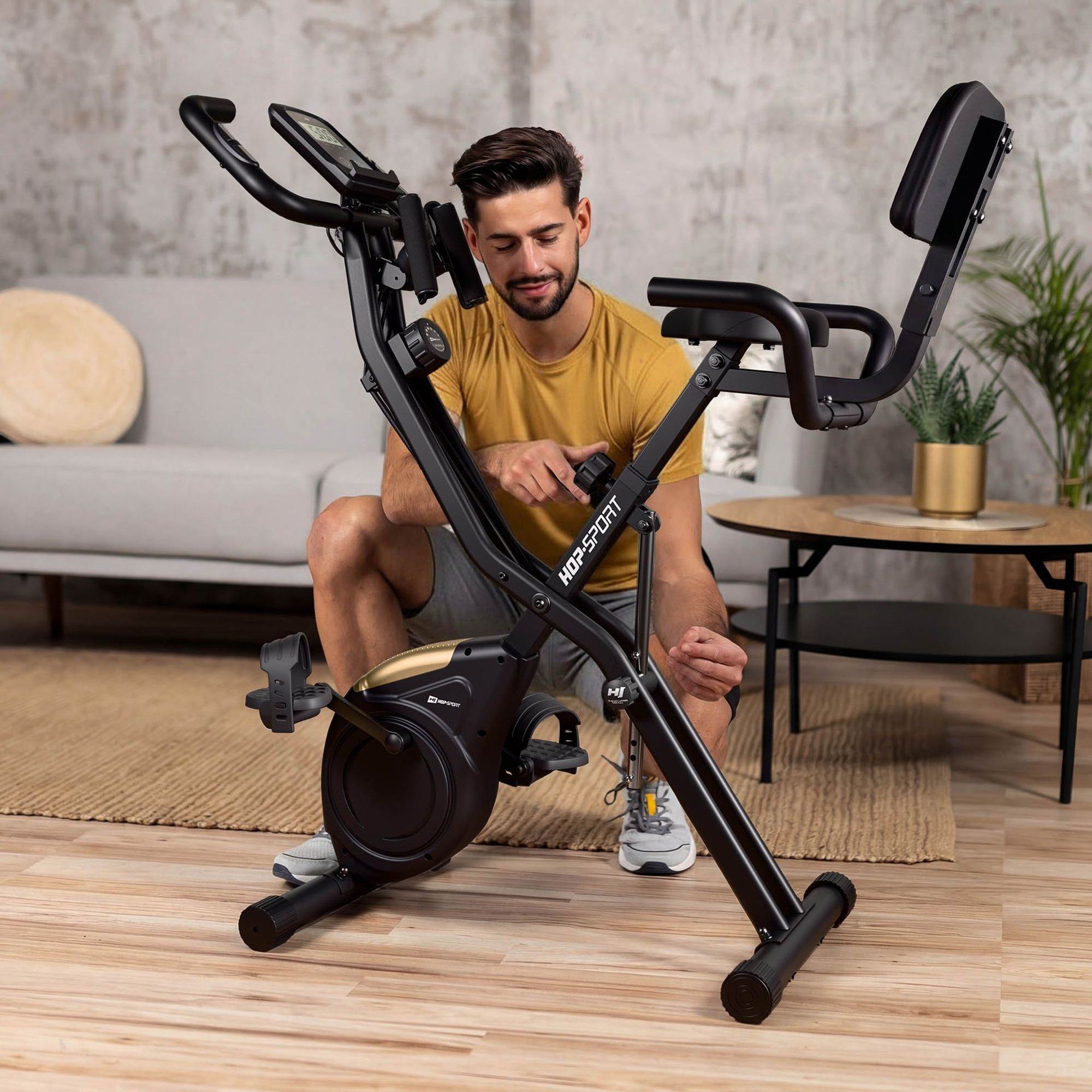 Ein Mann im gelben Shirt stellt drinnen ein schwarzes Heimfahrrad ein, mit Sofa und Pflanze im Hintergrund.
