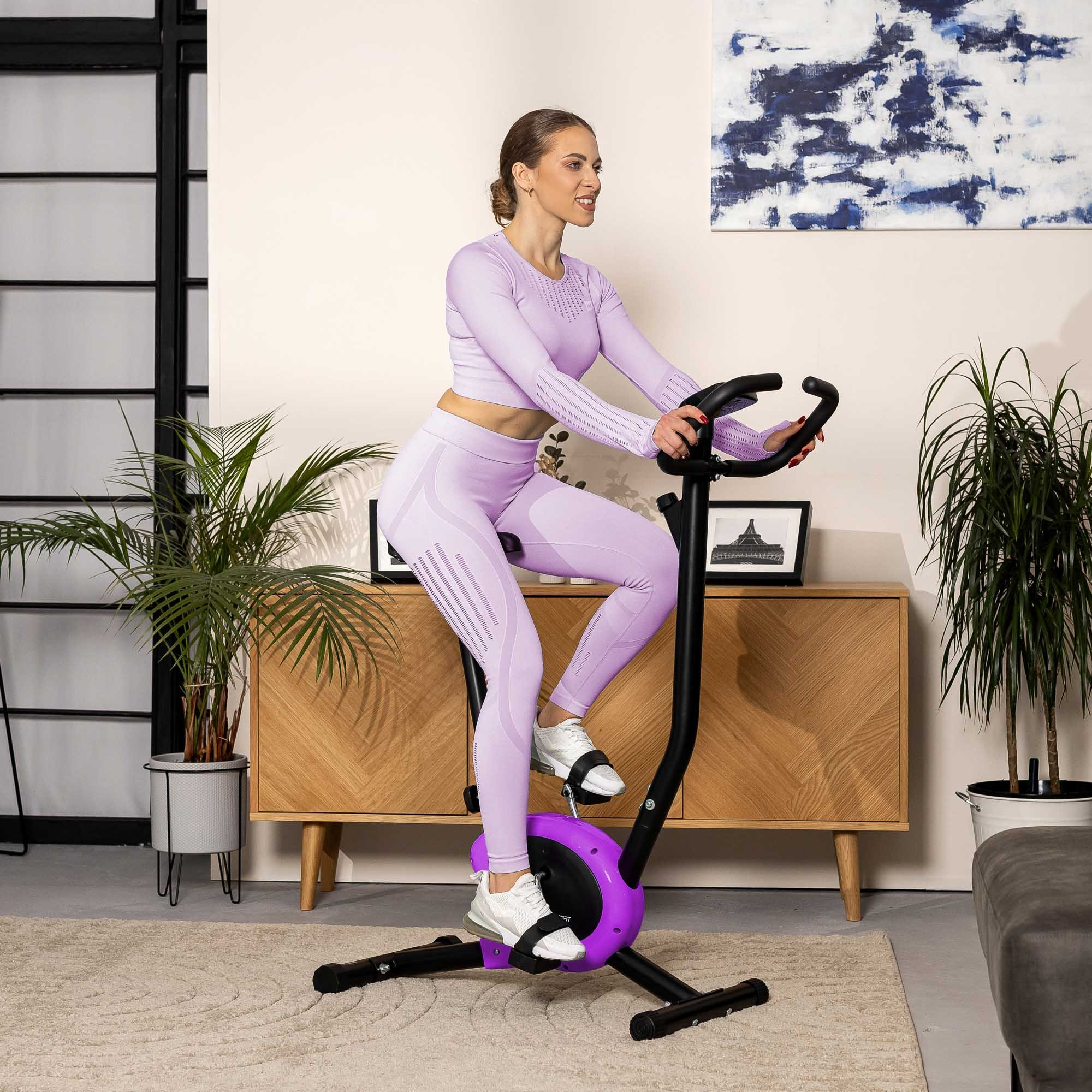 Eine Frau in einem hellvioletten Trainingsanzug fährt in einem hellen Wohnzimmer mit Pflanzen und einem Holzschrank Heimtrainer.