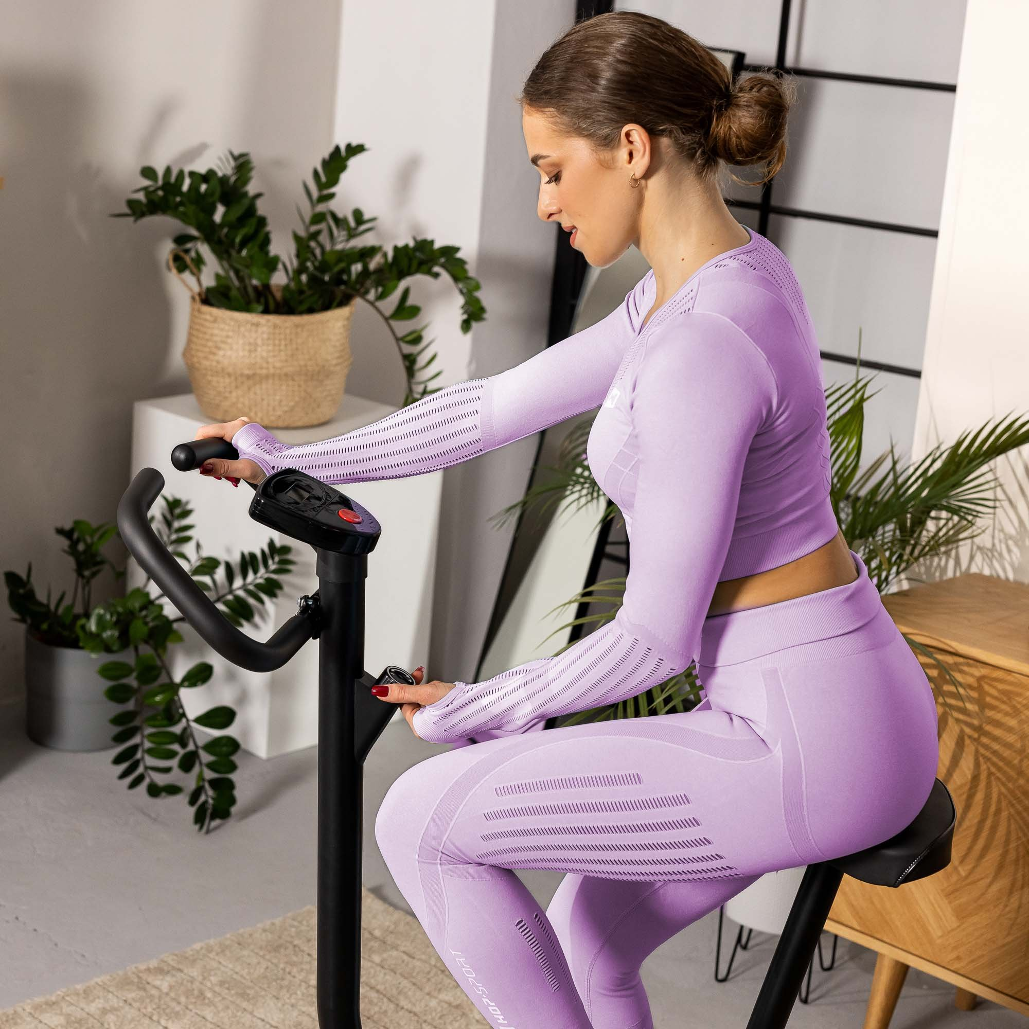 Eine Frau in einem hellvioletten Sportoutfit fährt in einem hellen Raum mit Topfpflanzen Heimtrainer.