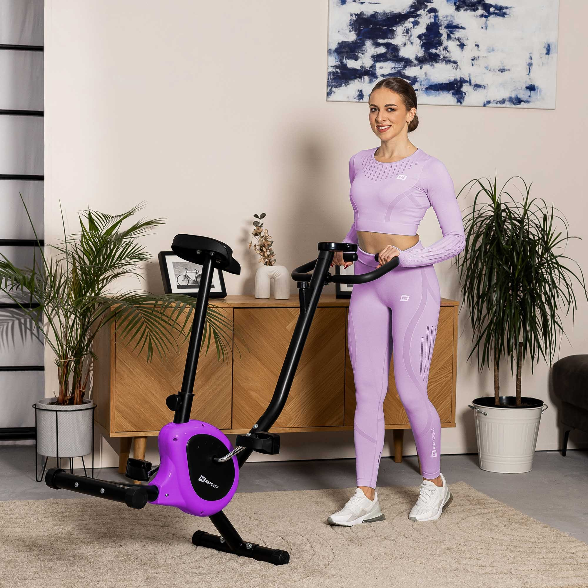 Eine Frau in lilafarbenem Sportoutfit steht im Wohnzimmer neben einem lila-schwarzen Heimtrainer.