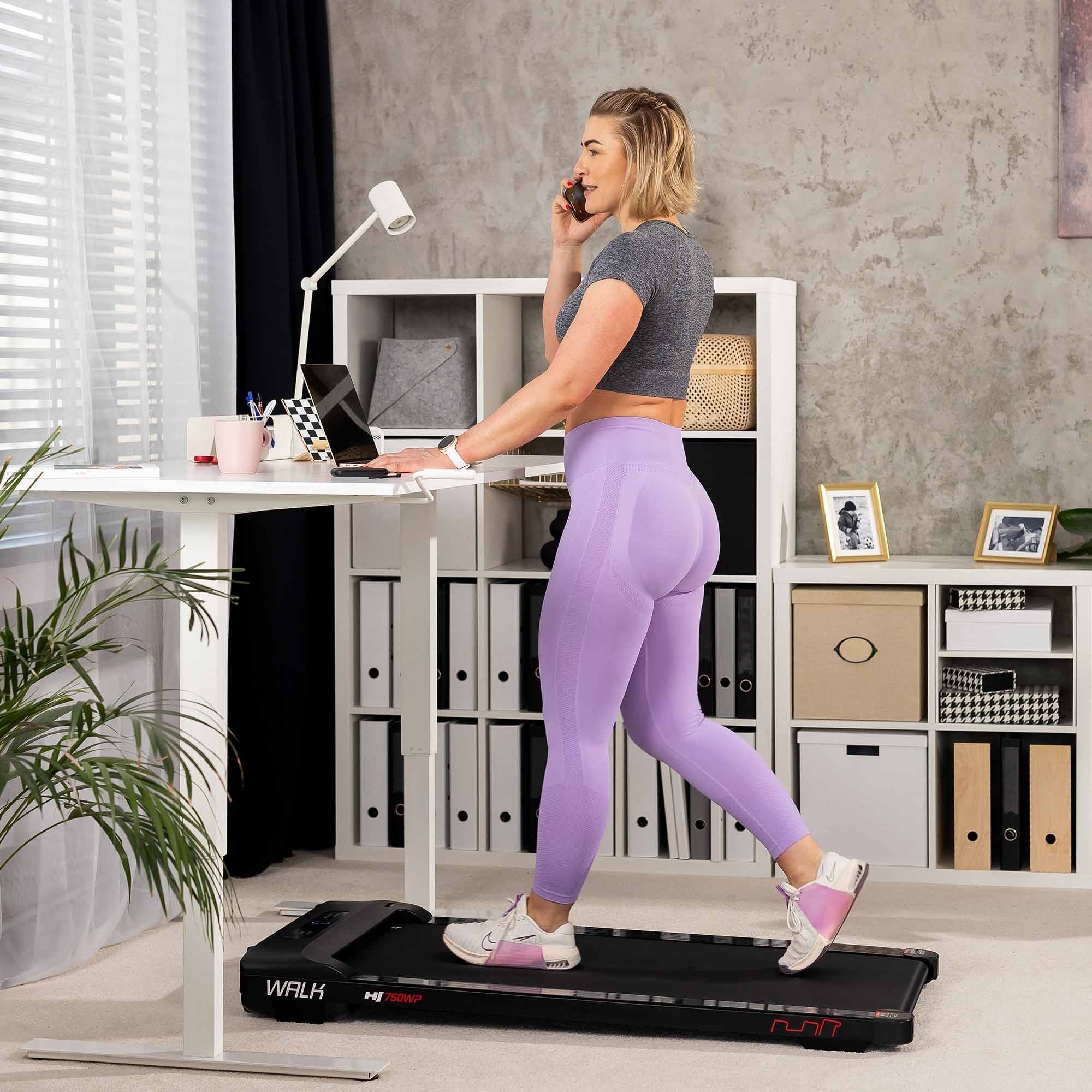 Eine Frau in lilafarbenen Leggings geht auf einem Untertisch-Laufband im Büro und telefoniert.