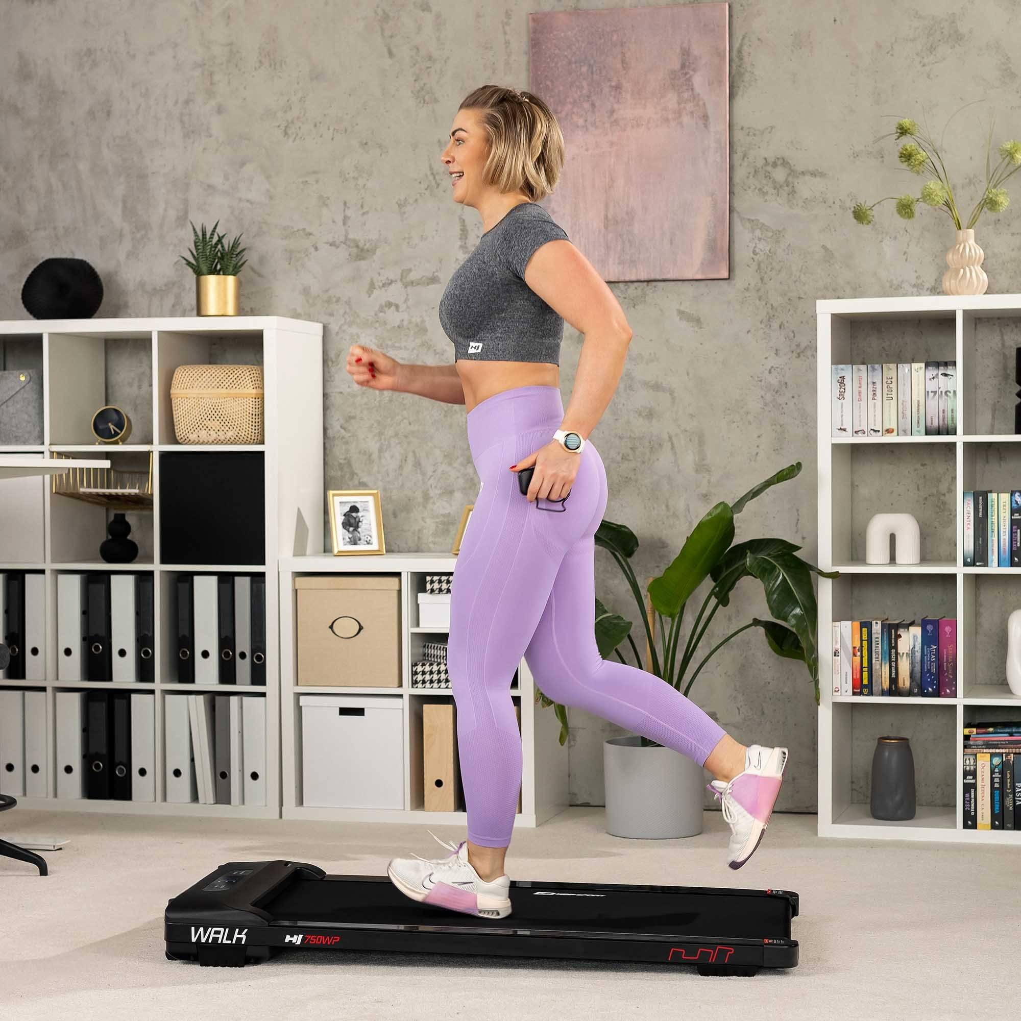 Eine blonde Frau in grauem Top und lila Leggings läuft auf einem schwarzen Laufband in einem Raum mit weißen Regalen.