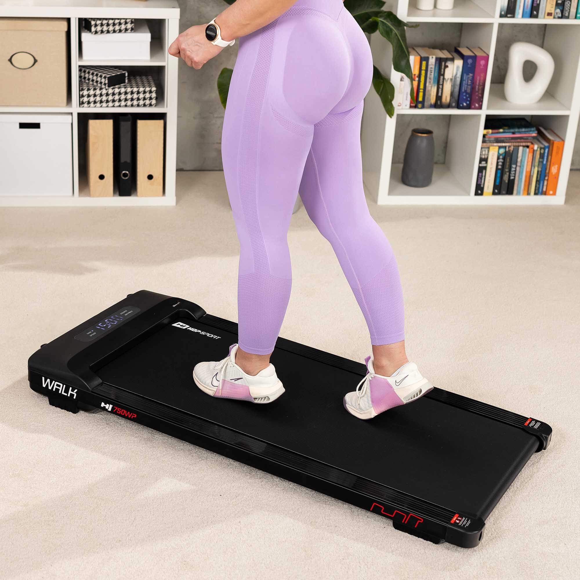 Eine Person in lila Leggings geht auf einem schwarzen Laufband, dahinter ein weißes Regal mit Büchern und Gegenständen.
