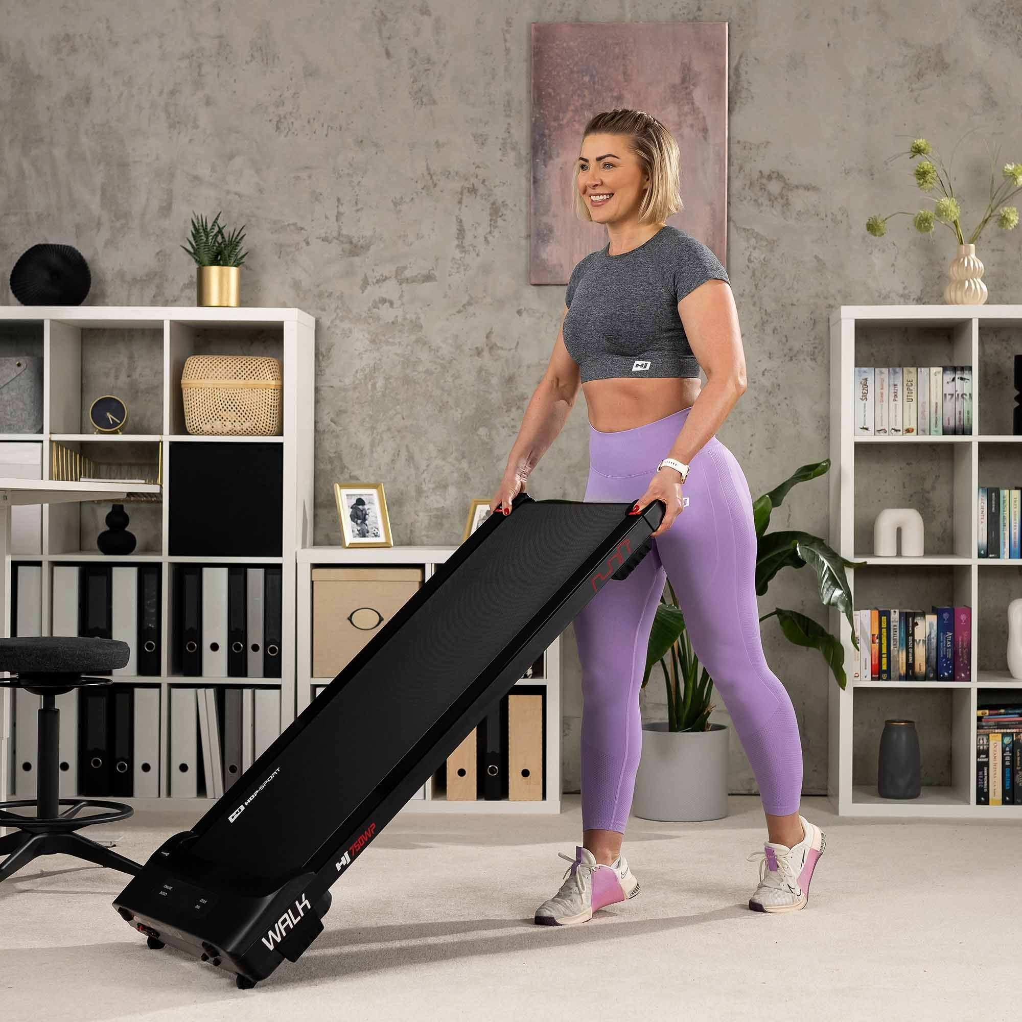 Eine lächelnde Frau in einem grauen Oberteil und lila Leggings kippt ein schwarzes Laufband in einem hellen Raum.