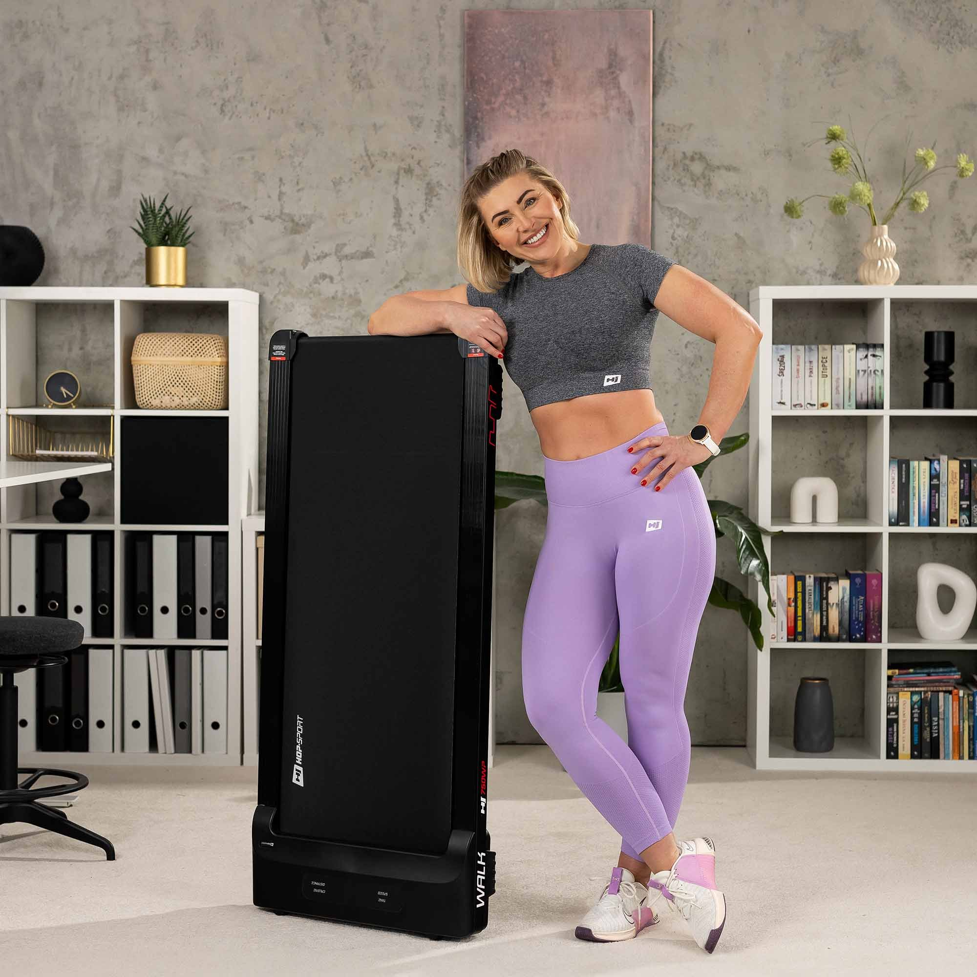 Lächelnende Frau in Sportbekleidung stützt sich in einem hellen Raum auf ein zusammengeklapptes Laufband.