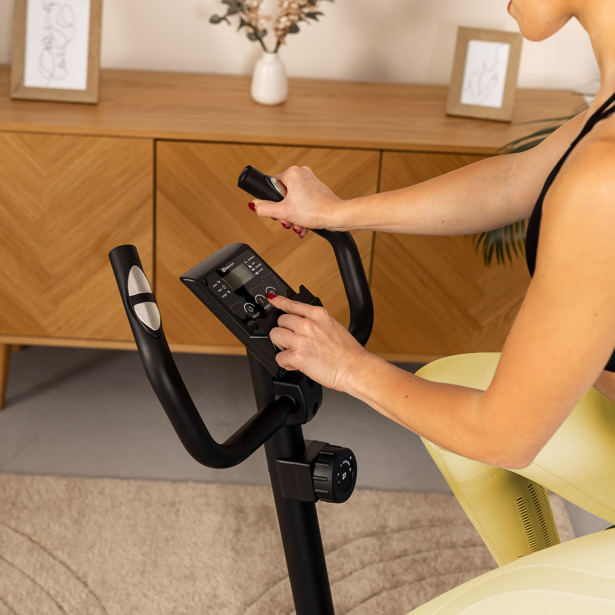 Eine Frau in gelben Leggings stellt den Trainingscomputer eines Heimtrainers in einem Wohnzimmer ein.
