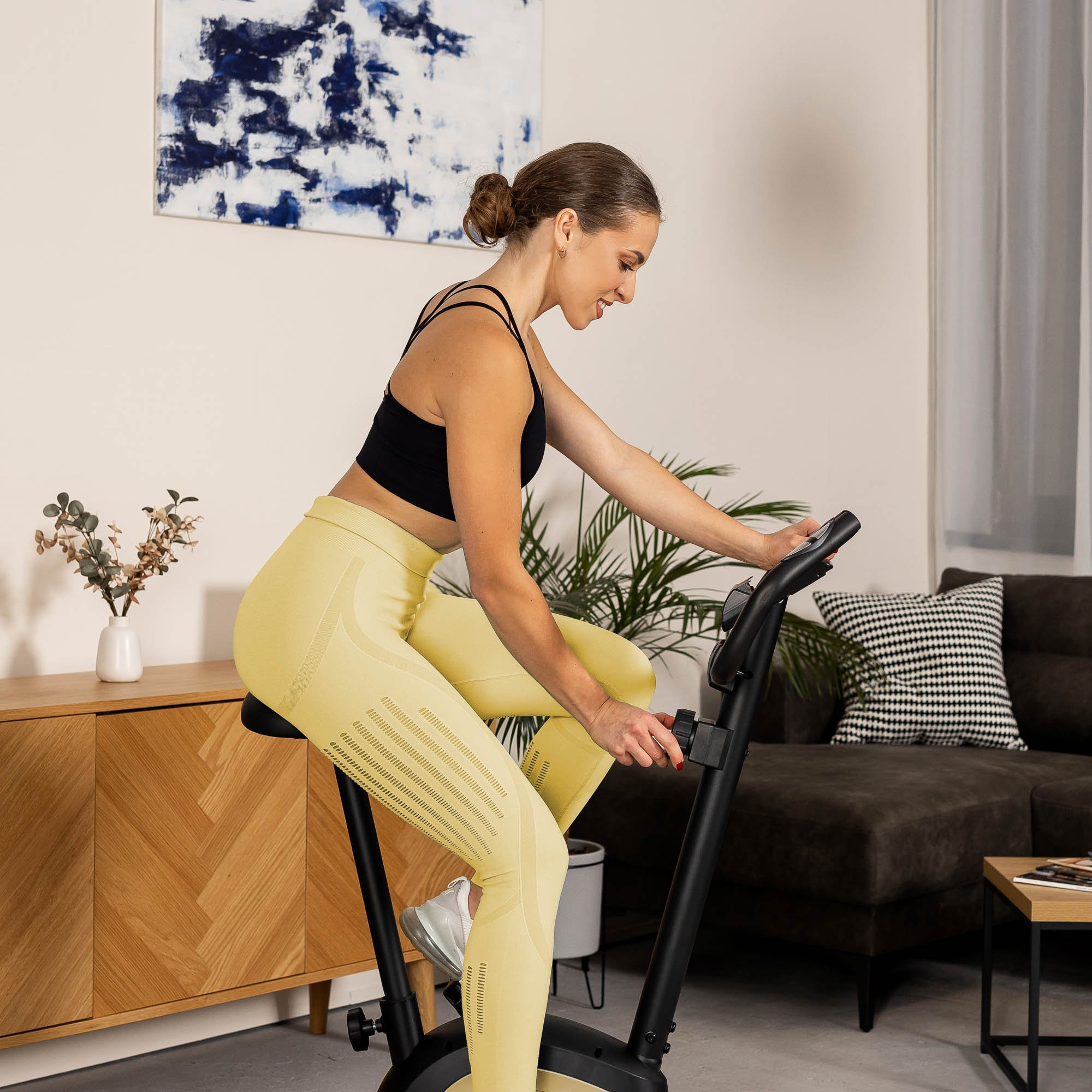 Eine Frau in gelben Leggings und schwarzem Top trainiert auf einem Heimtrainer in einem hellen Zimmer.