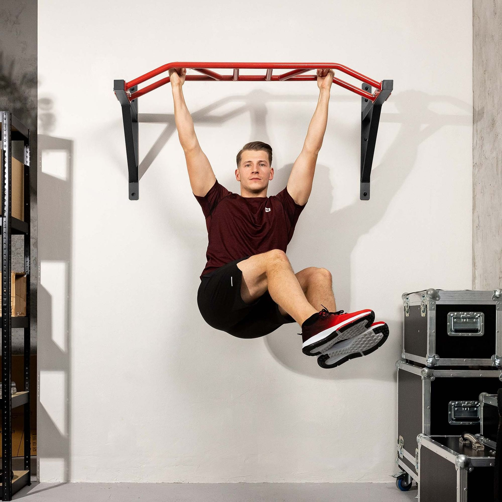 Ein Mann in Sportkleidung hängt an einer roten Wandklimmzugstange und hebt die Beine an.