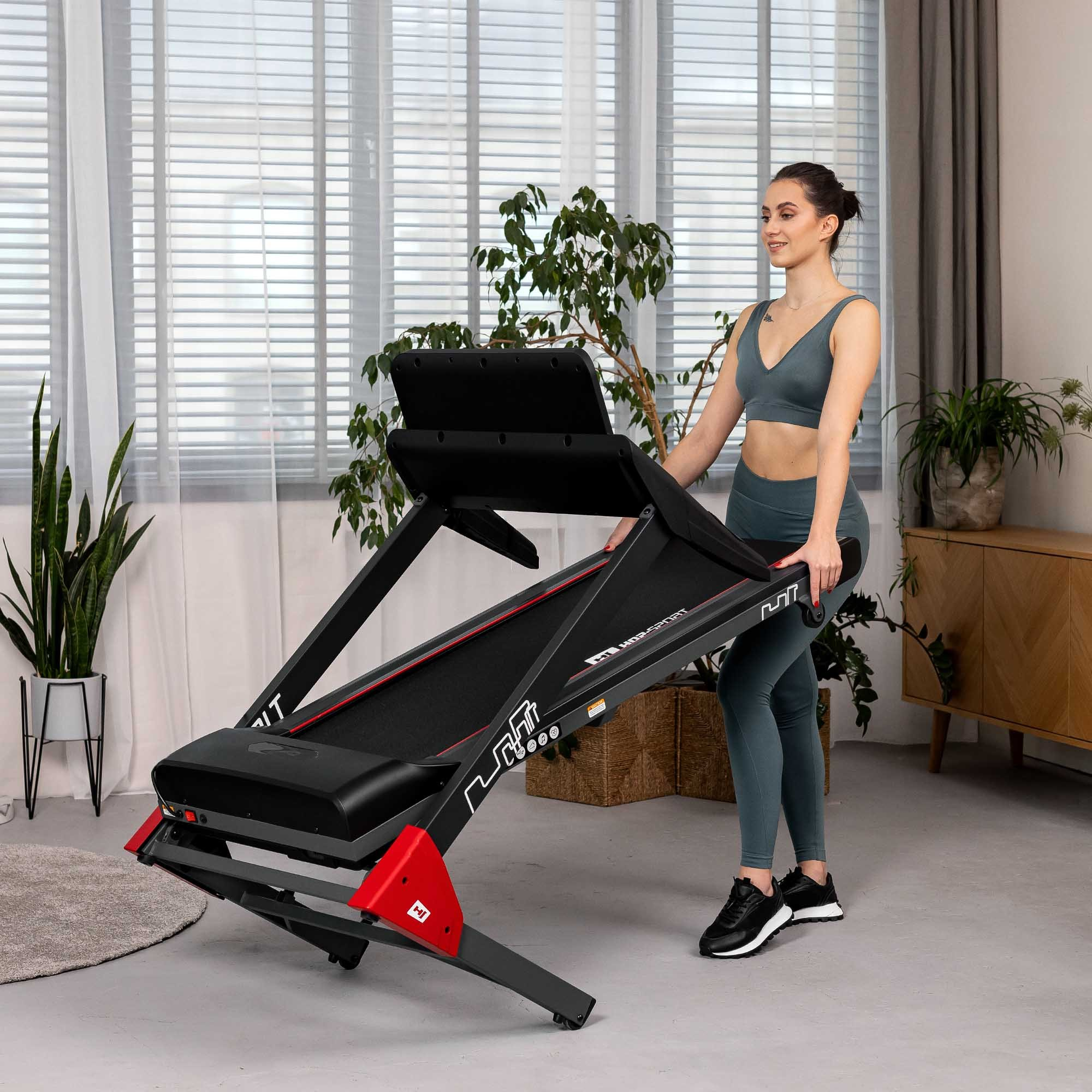 Frau in Sportkleidung klappt ein schwarzes und rotes Laufband in einem hellen Raum zusammen.