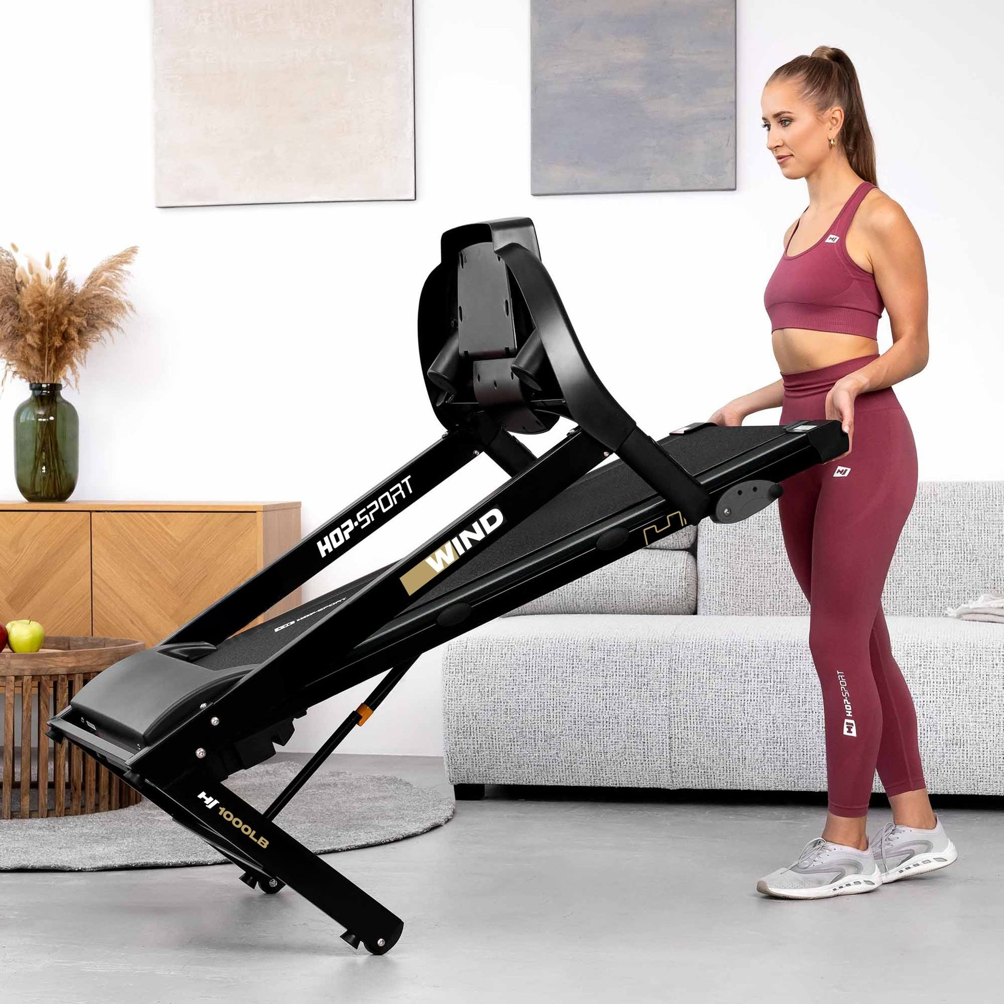 Eine Frau im passenden rosa Sportoutfit klappt drinnen ein schwarzes Hop-Sport Wind Laufband zusammen.