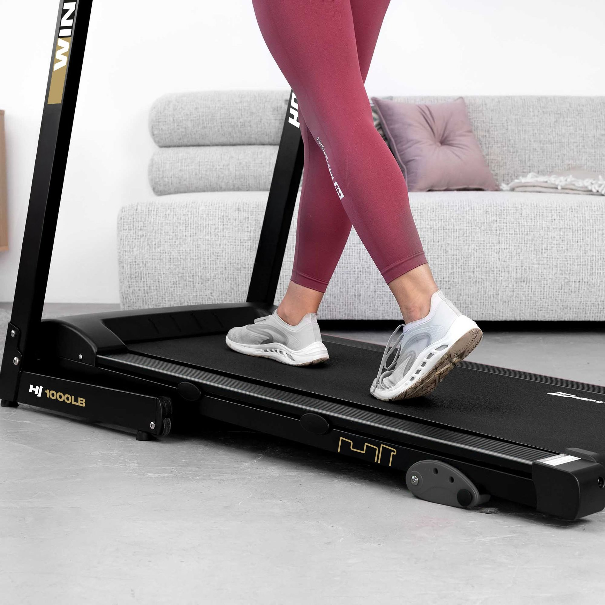 Person in roten Leggings geht auf einem schwarzen Laufband vor einem hellen Sofa.