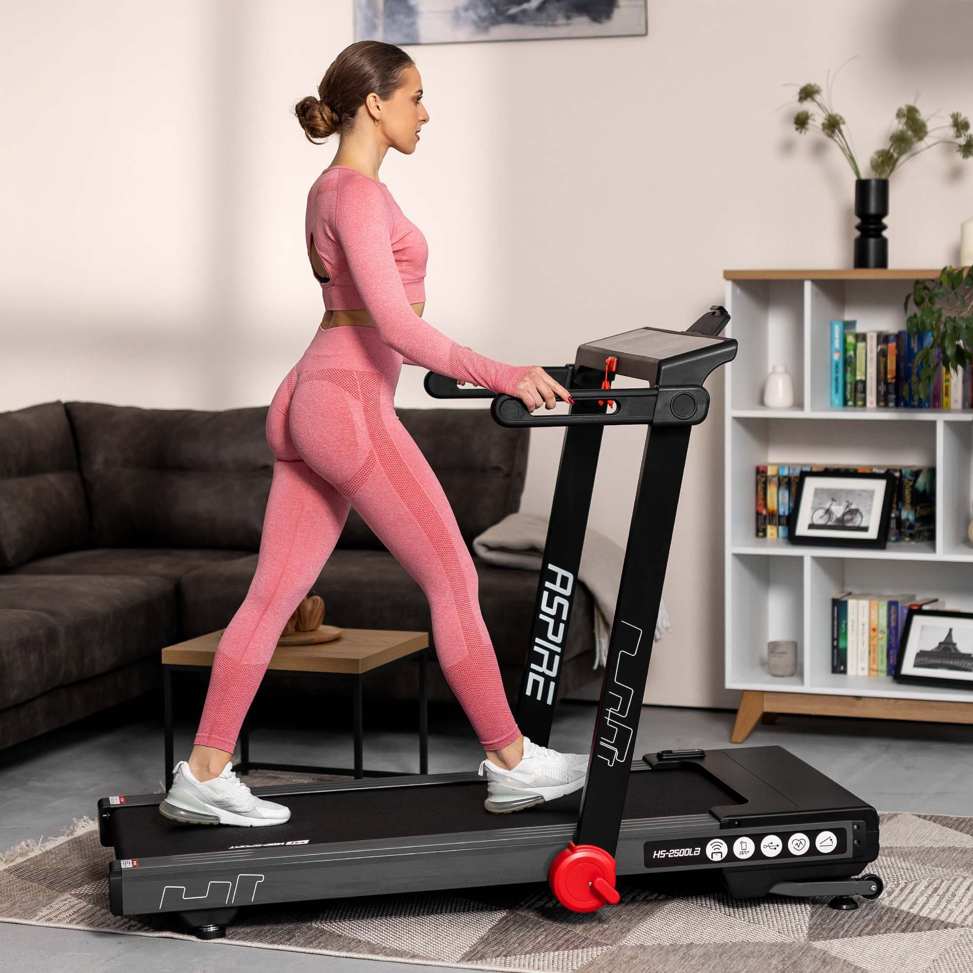 Eine Frau in pinker Sportkleidung geht auf einem schwarzen Laufband in einem gemütlichen Raum.