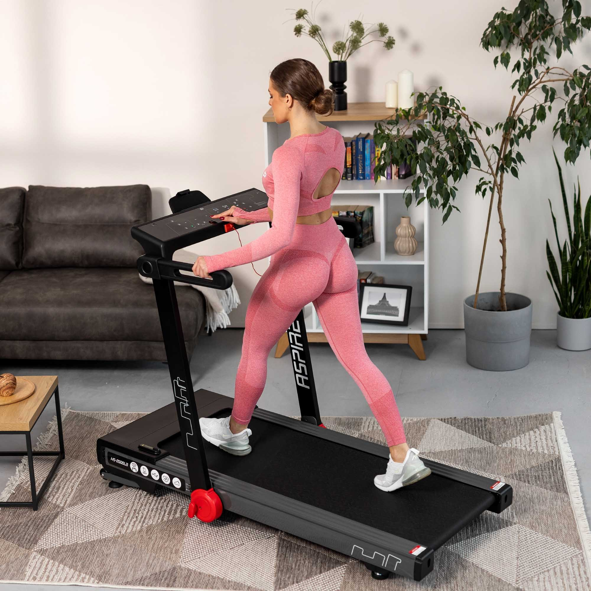Eine Frau in pinker Sportkleidung trainiert zu Hause auf einem schwarzen Laufband vor einem hellen Regal und einer Couch.