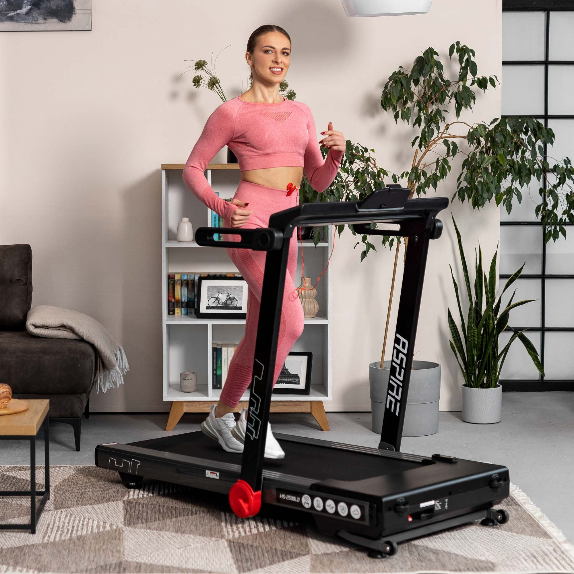 Eine Frau in pinker Sportkleidung läuft auf einem schwarzen Laufband in einem hellen Wohnzimmer mit Pflanzen.