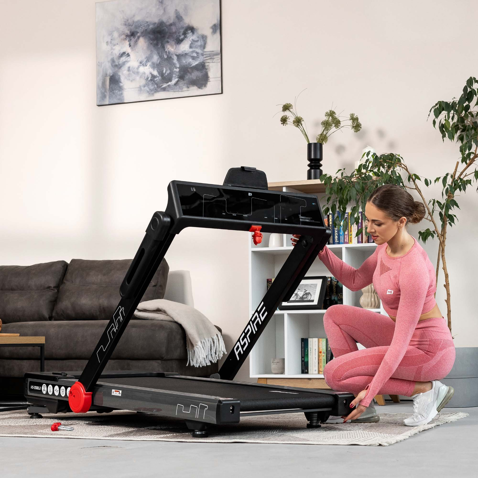 Eine Frau in rosa Sportkleidung kniet, um ein schwarzes Aspire-Laufband zusammenzuklappen.