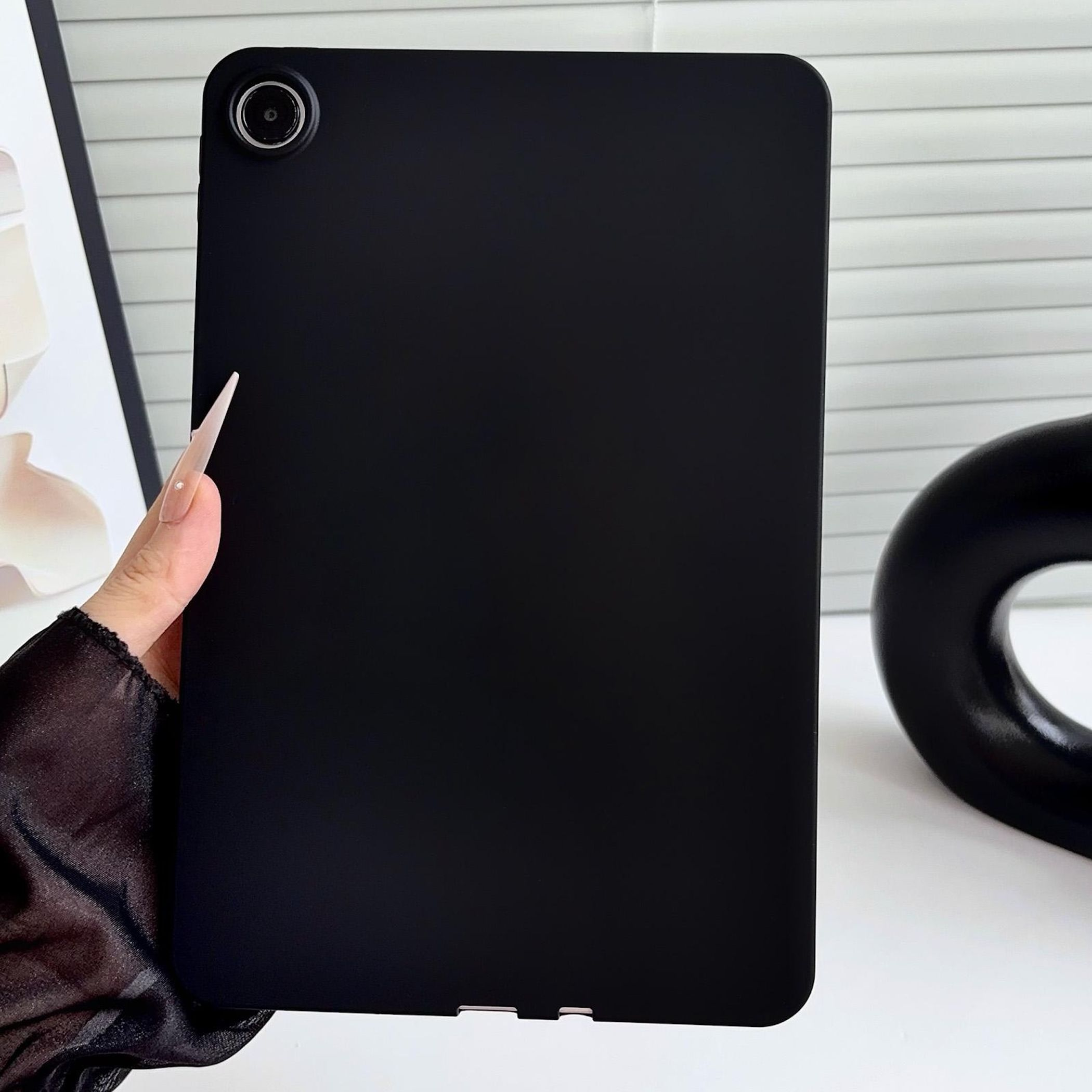 Una mano sostiene una custodia nera per tablet in verticale contro una superficie bianca con un oggetto decorativo.