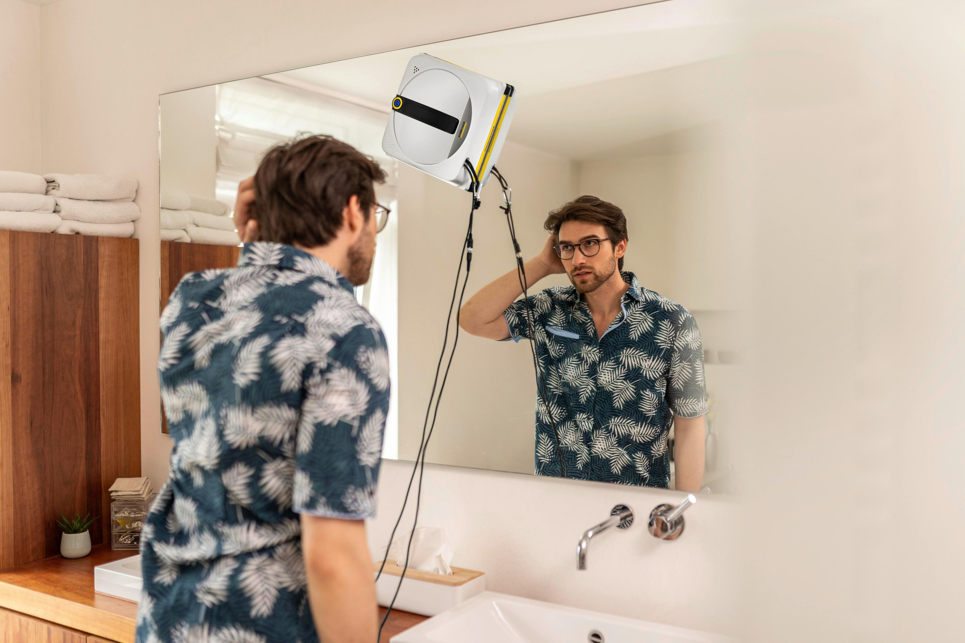 Un uomo con una camicia blu si guarda nello specchio del bagno mentre un robot puliscivetri pulisce il centro.