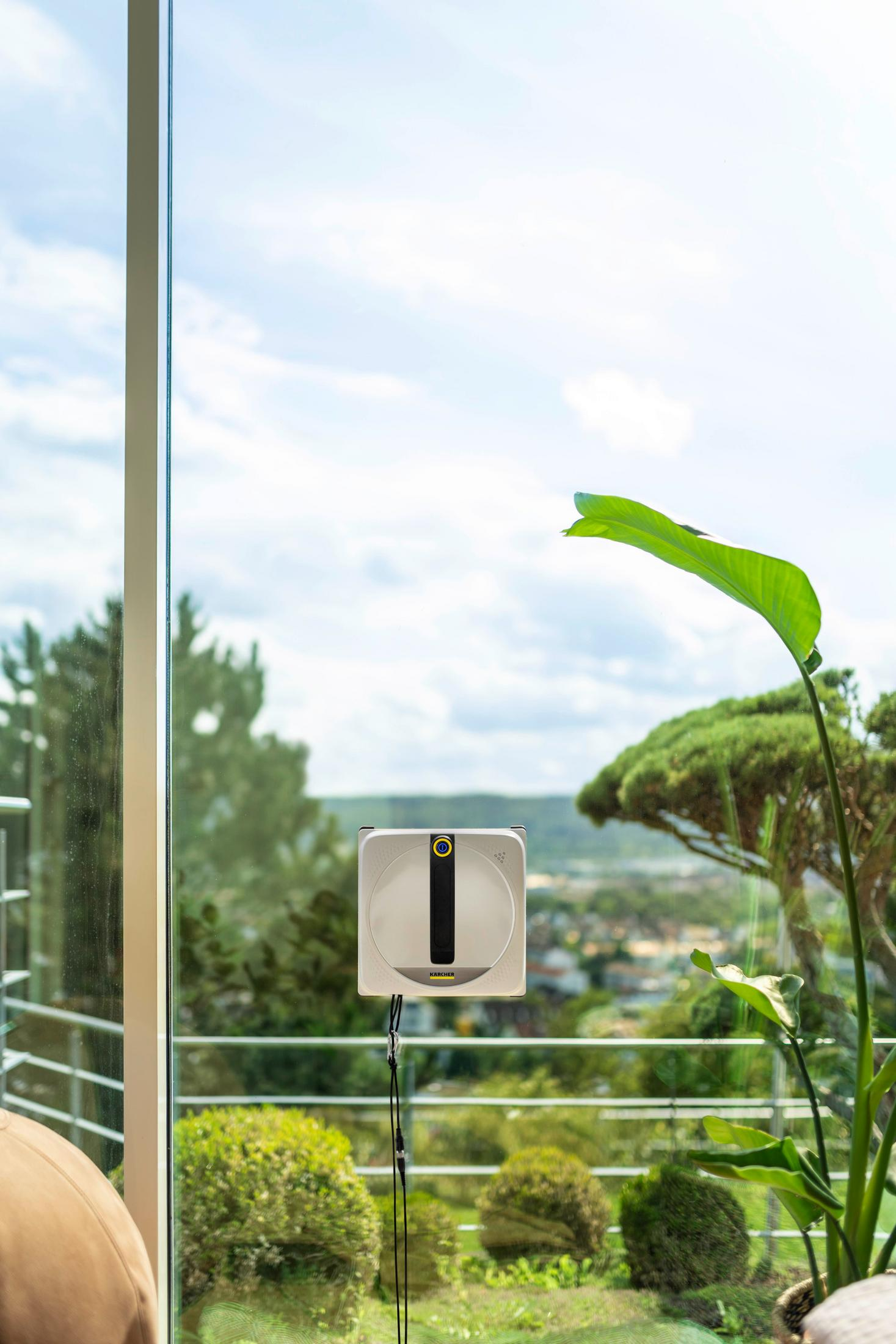 Un robot pulisci vetri bianco è attaccato a una grande vetrata, con alberi e un cielo nuvoloso sullo sfondo.