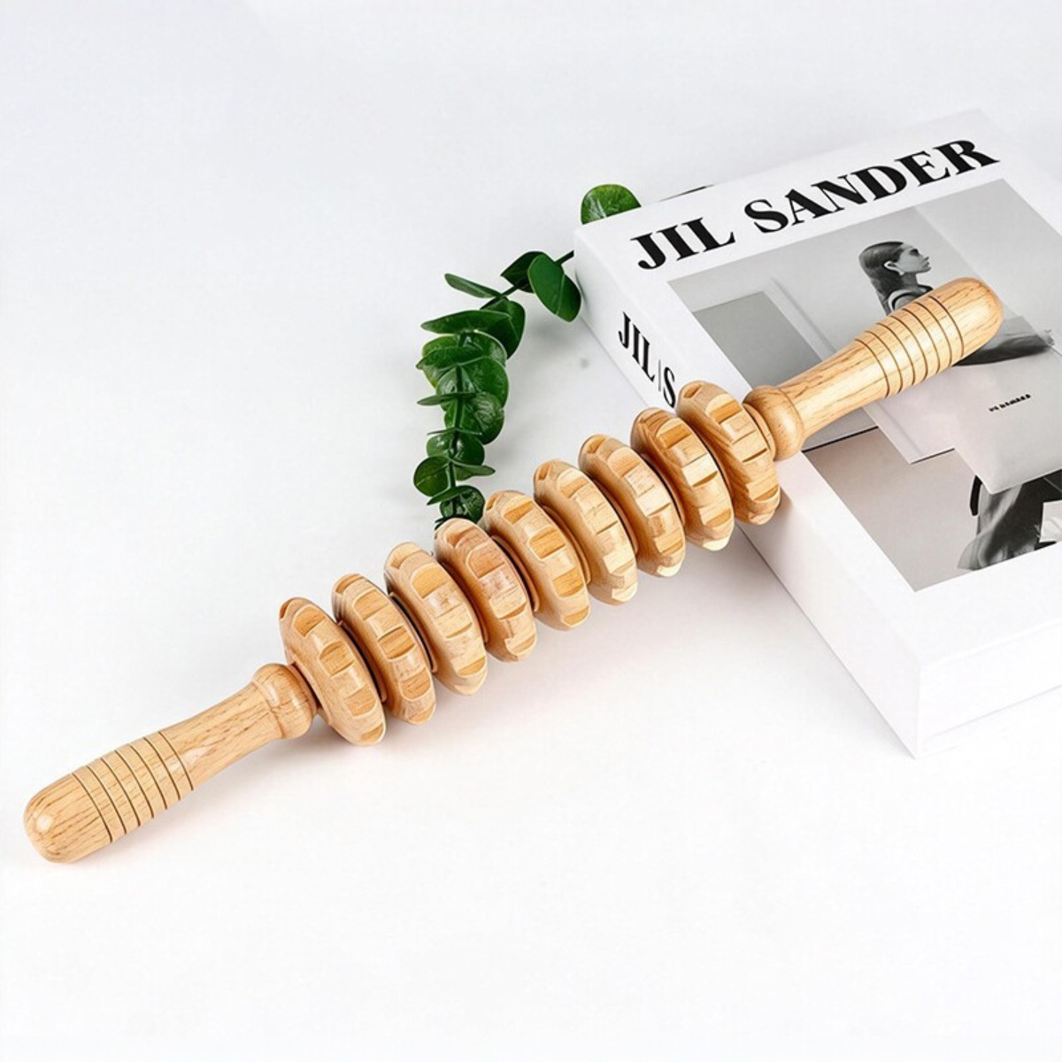 Ein hölzerner Massagestab mit geriffelten Rädern liegt neben einem Jil Sander Buch und Eukalyptuszweigen.