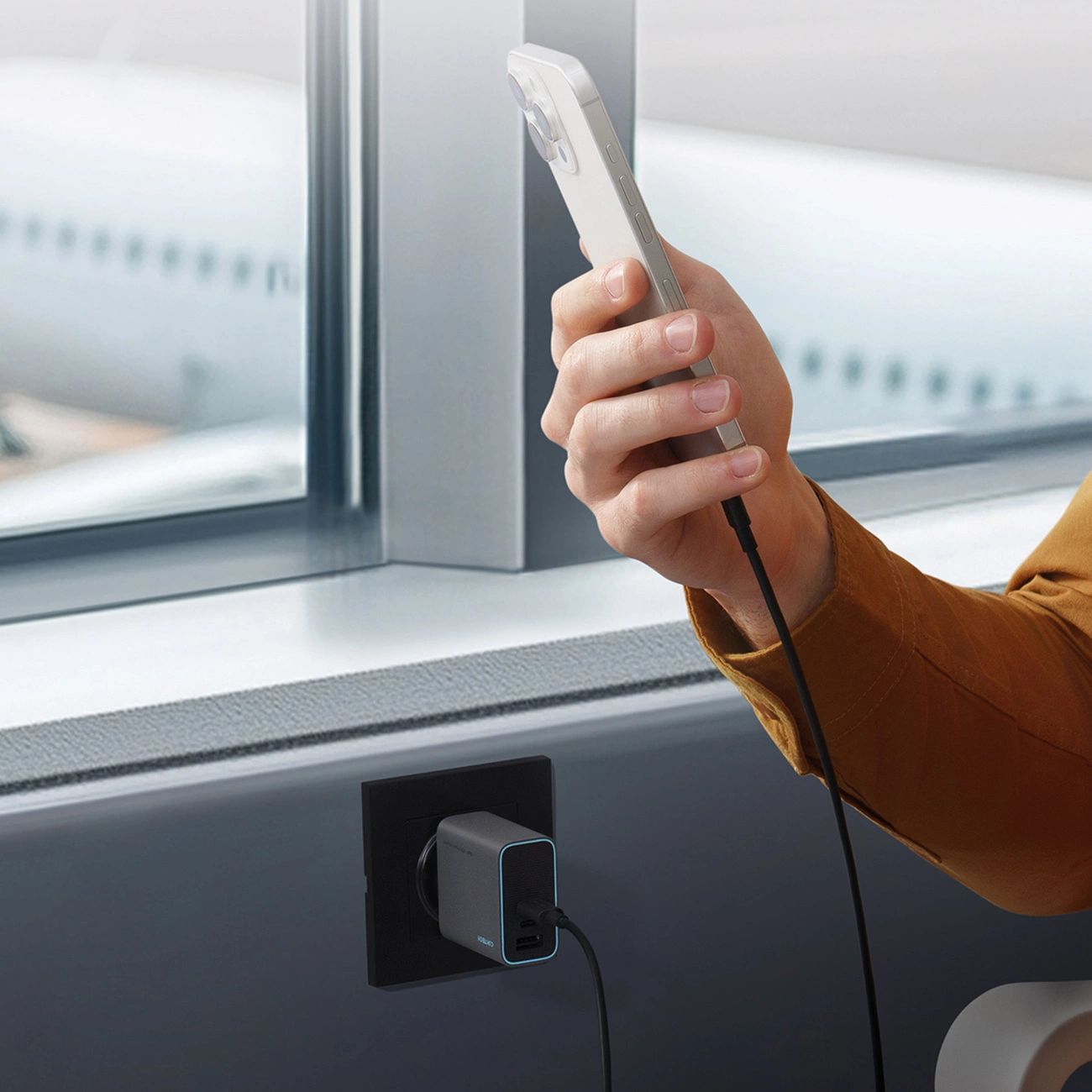 Eine Person hält ein Handy, das per Kabel mit einem grauen Ladegerät in einer Steckdose verbunden ist.