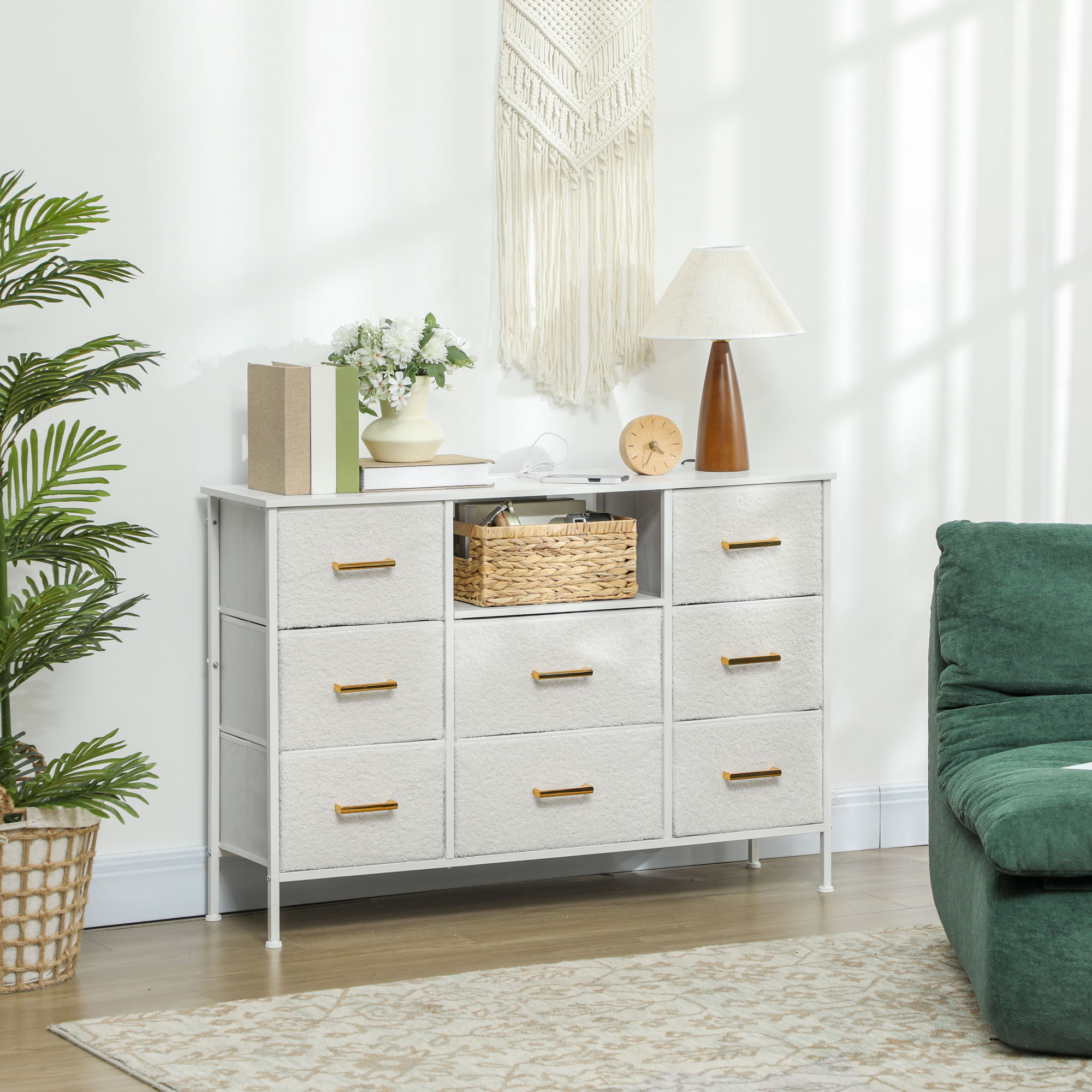 Una cómoda blanca con cajones de tela contra una pared blanca, decorada con una lámpara y flores.
