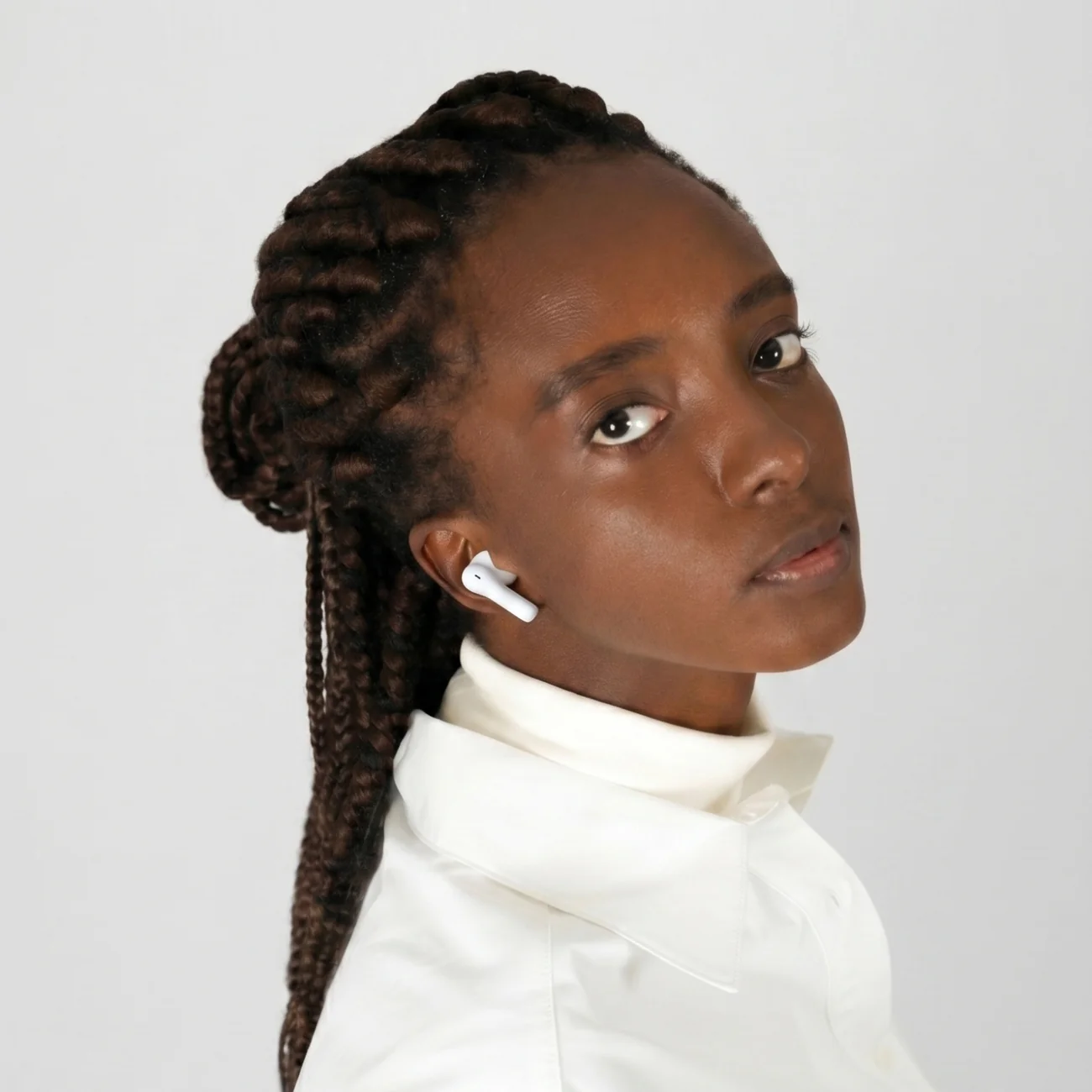 Una joven con trenzas marrones y auriculares blancos lleva una camisa blanca sobre un fondo claro.