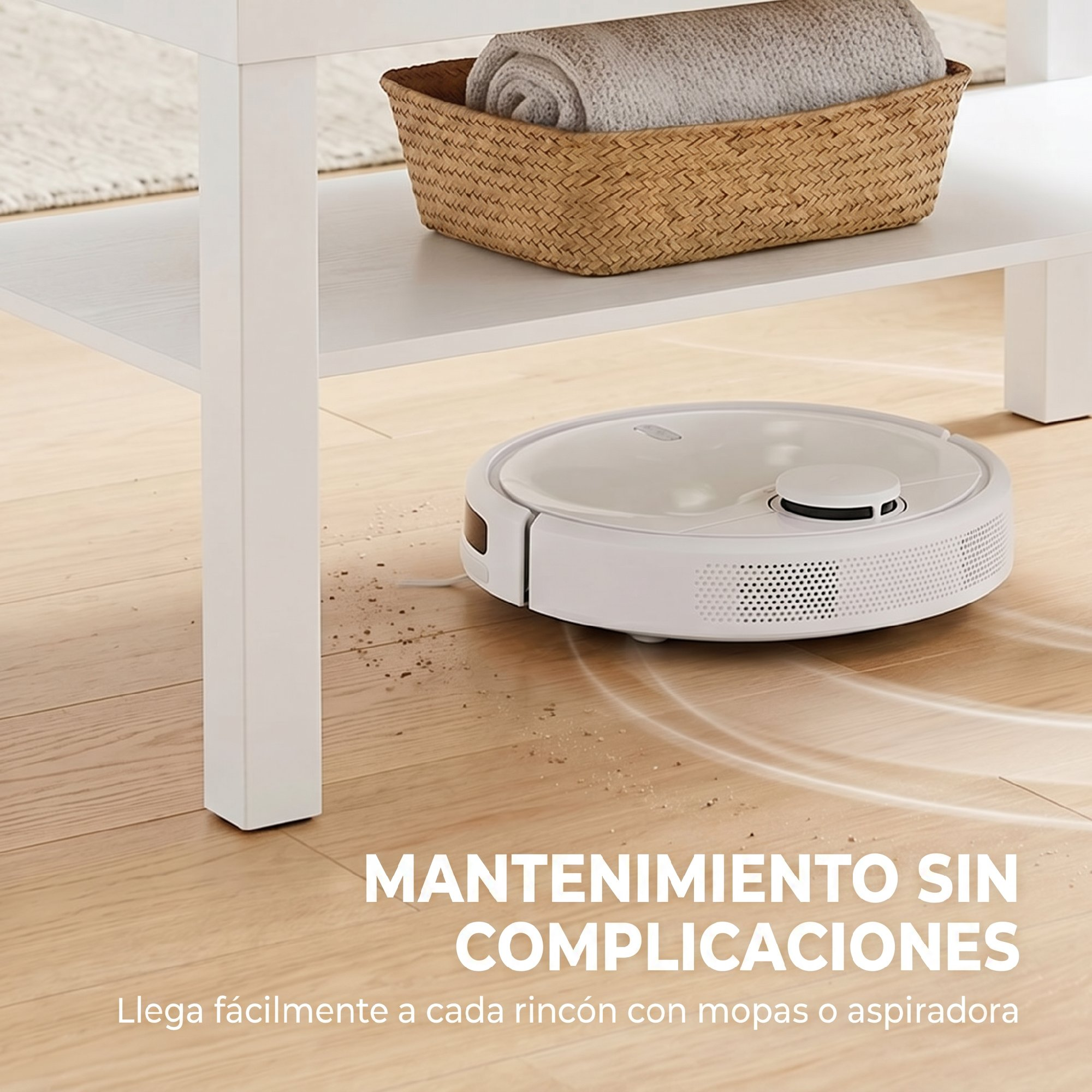 Un robot aspirador blanco limpia un suelo de madera bajo una mesa blanca con una cesta de mimbre y una manta.