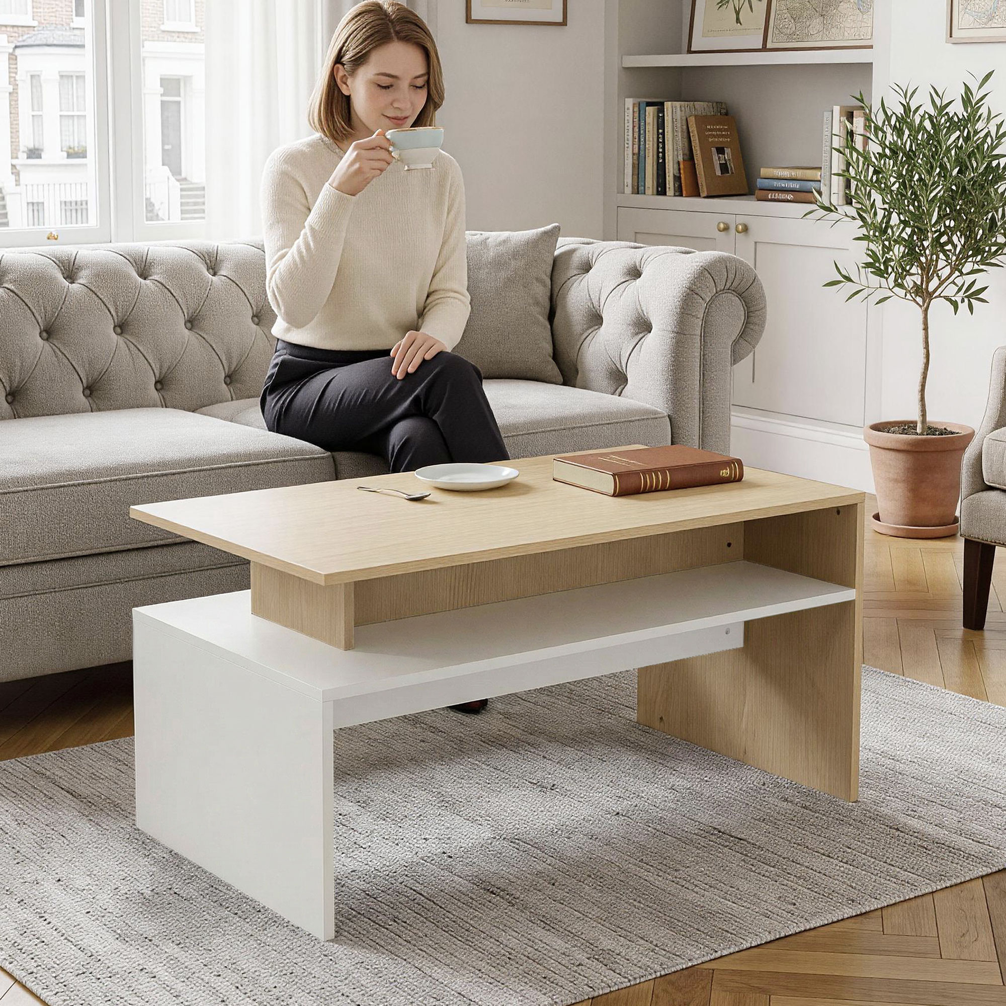 Una mujer está sentada en un sofá gris bebiendo de una taza, con una mesa de centro moderna de madera y blanca en primer plano.