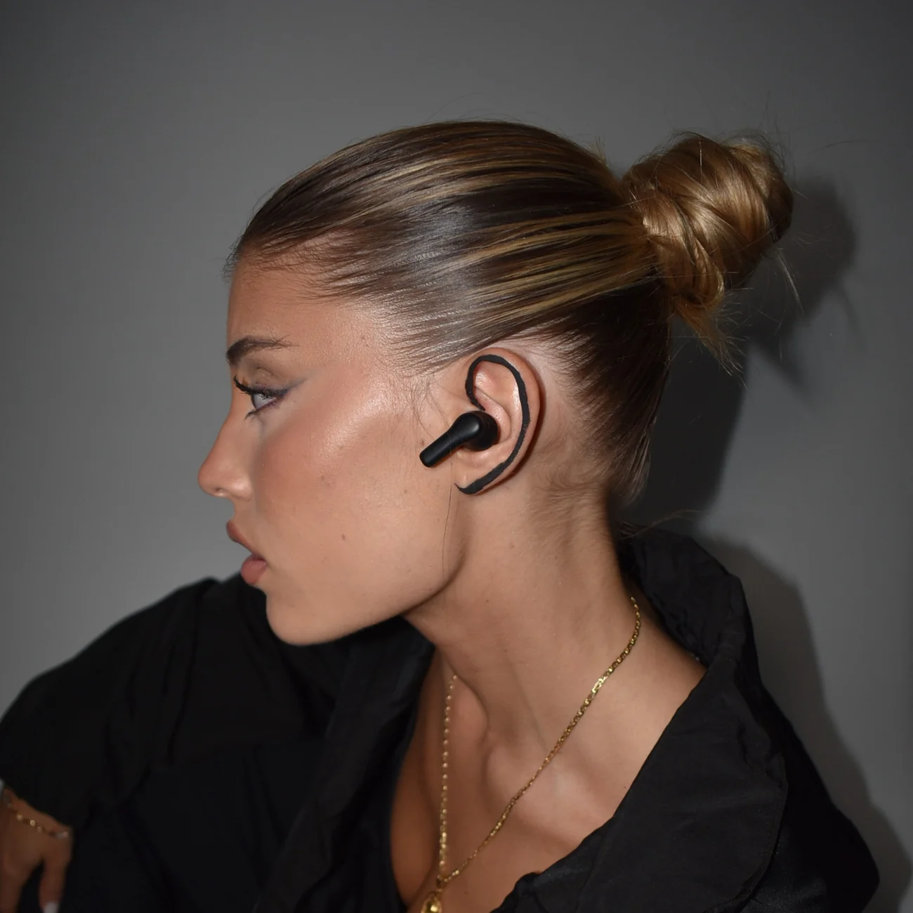 Perfil de una mujer con cabello rubio en moño con un auricular inalámbrico negro y collar de oro contra una pared gris.