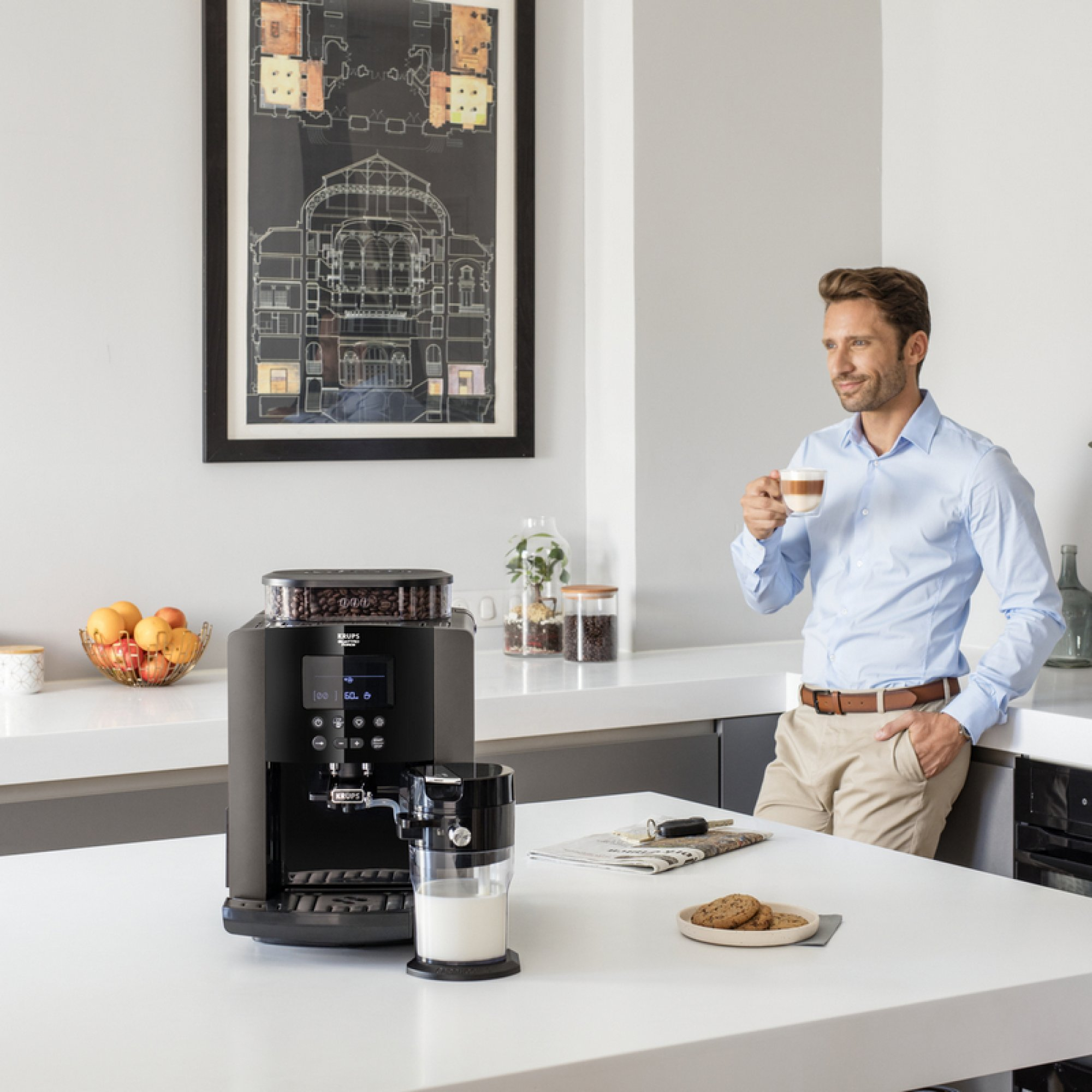 Un hombre con camisa azul sostiene una taza de café en una cocina moderna con una cafetera negra.