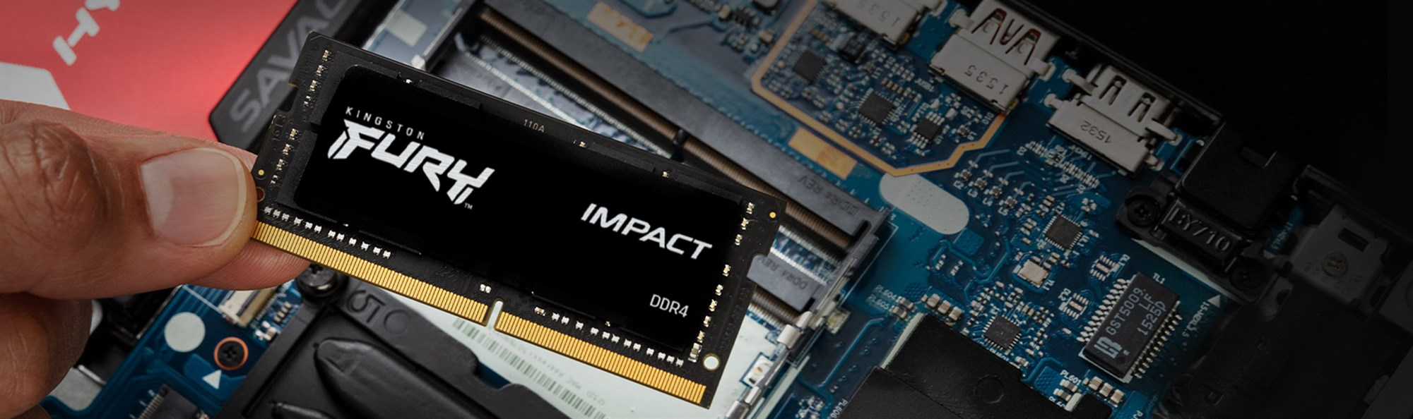 A hand holds a Kingston Fury Impact DDR4 RAM stick above a blue laptop motherboard.