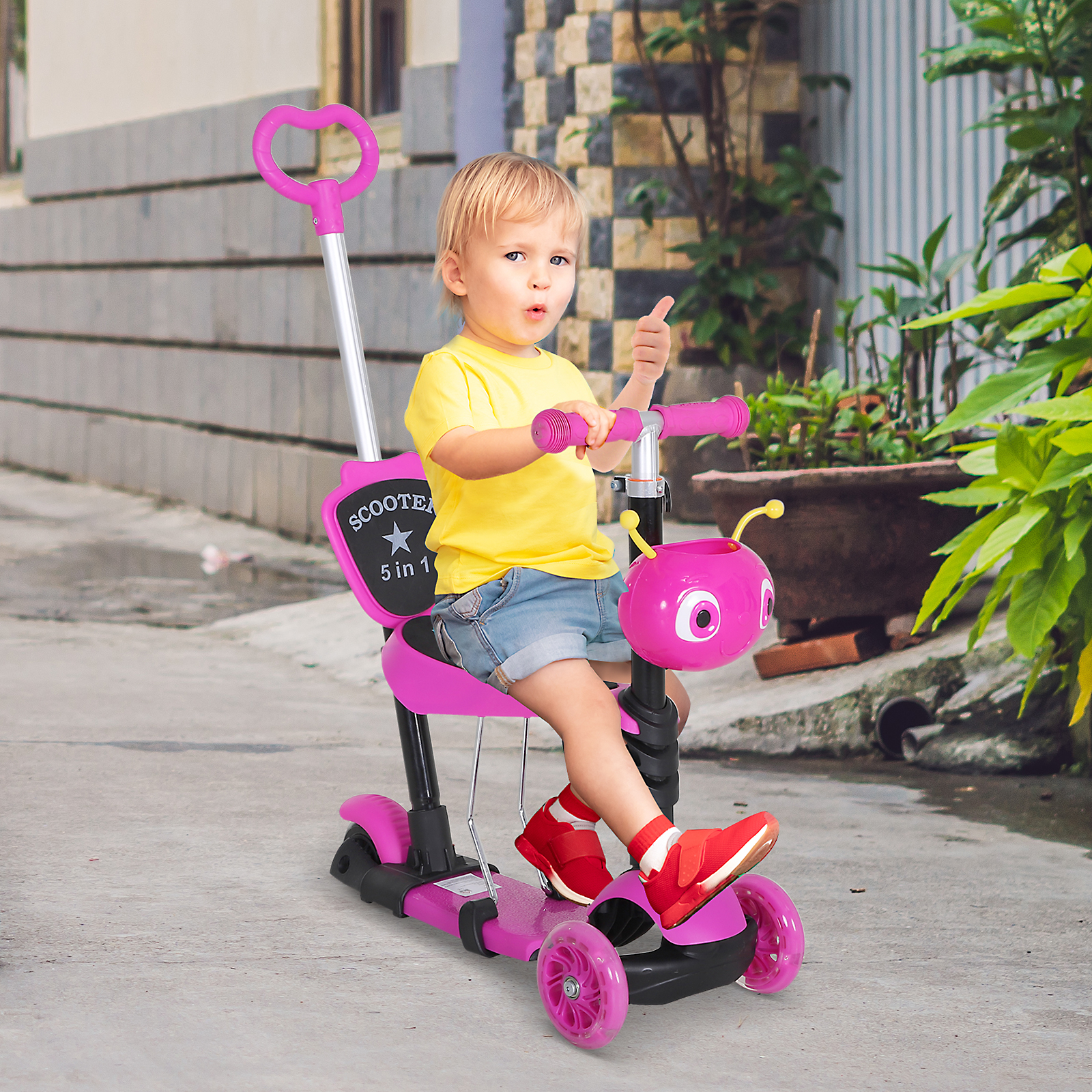 Bambino con maglietta gialla su un monopattino rosa 5 in 1 su un marciapiede, fa il pollice in su.