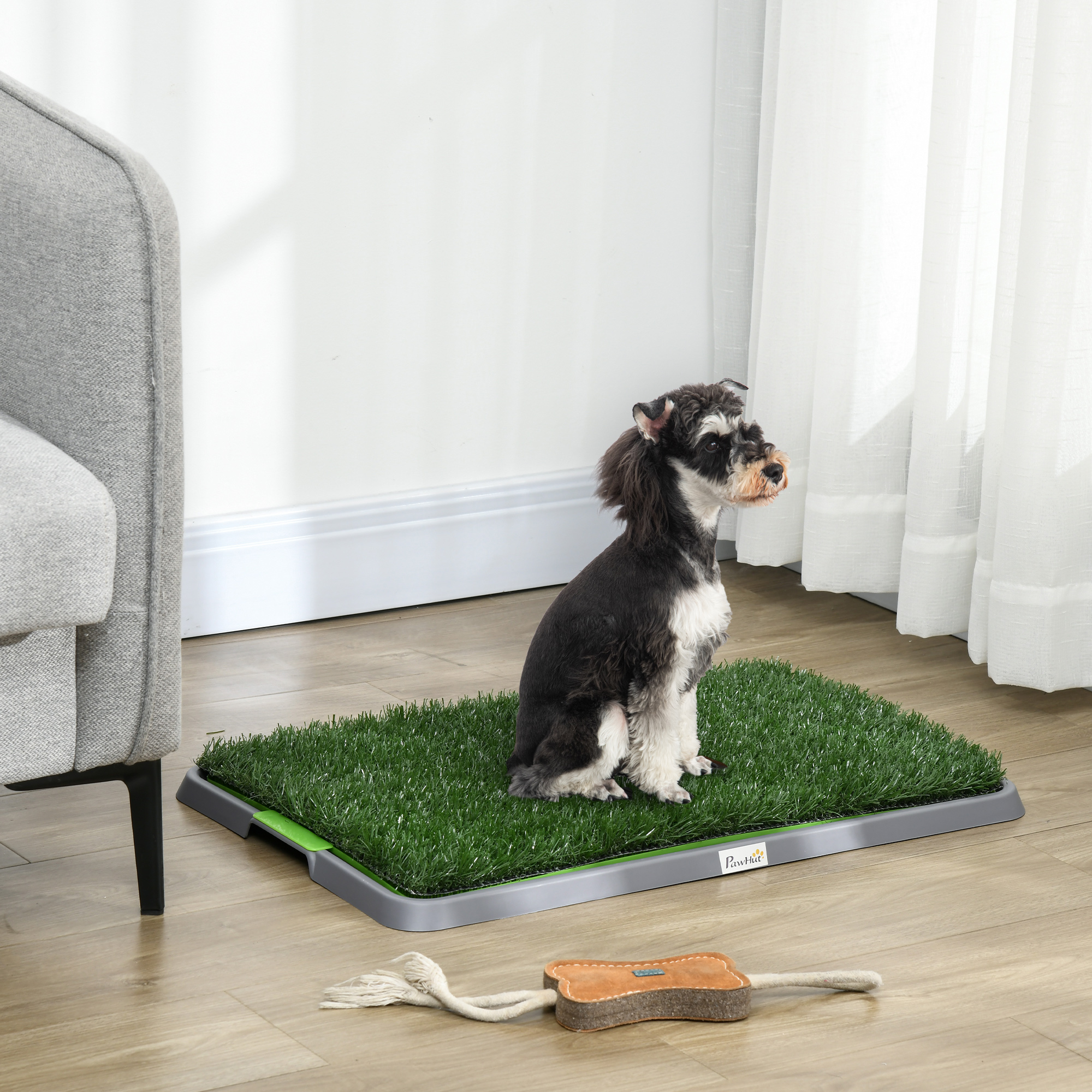 Un cane bianco e nero siede su un prato sintetico per cani su un pavimento in legno vicino a una poltrona grigia.