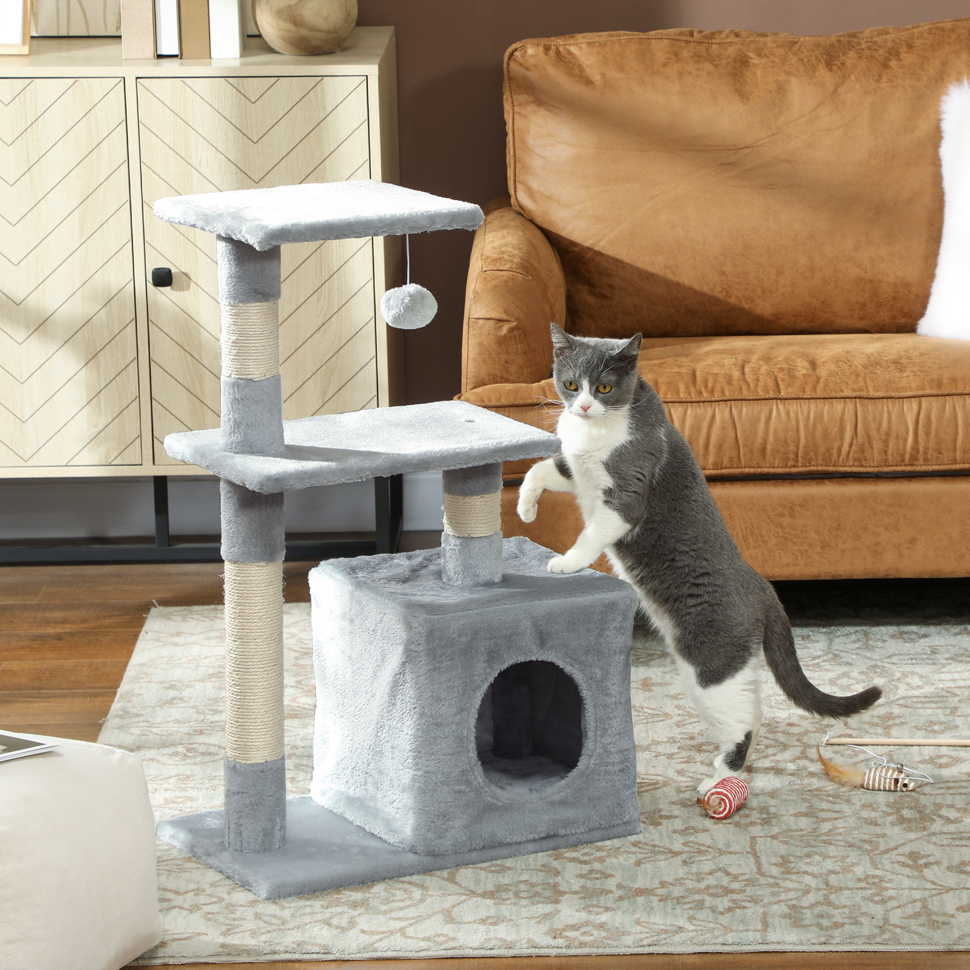 Un gatto grigio e bianco si trova accanto a un tiragraffi grigio a più livelli su un tappeto davanti a un divano marrone.