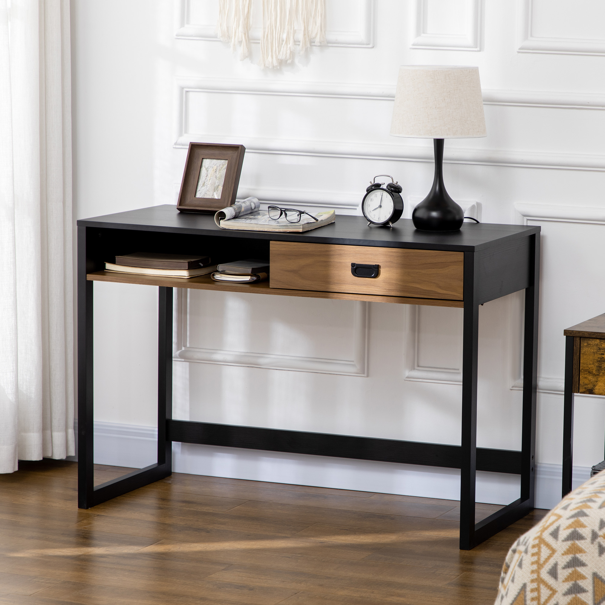 Una scrivania nera con cassetto in legno, lampada, orologio e cornice contro una parete bianca.