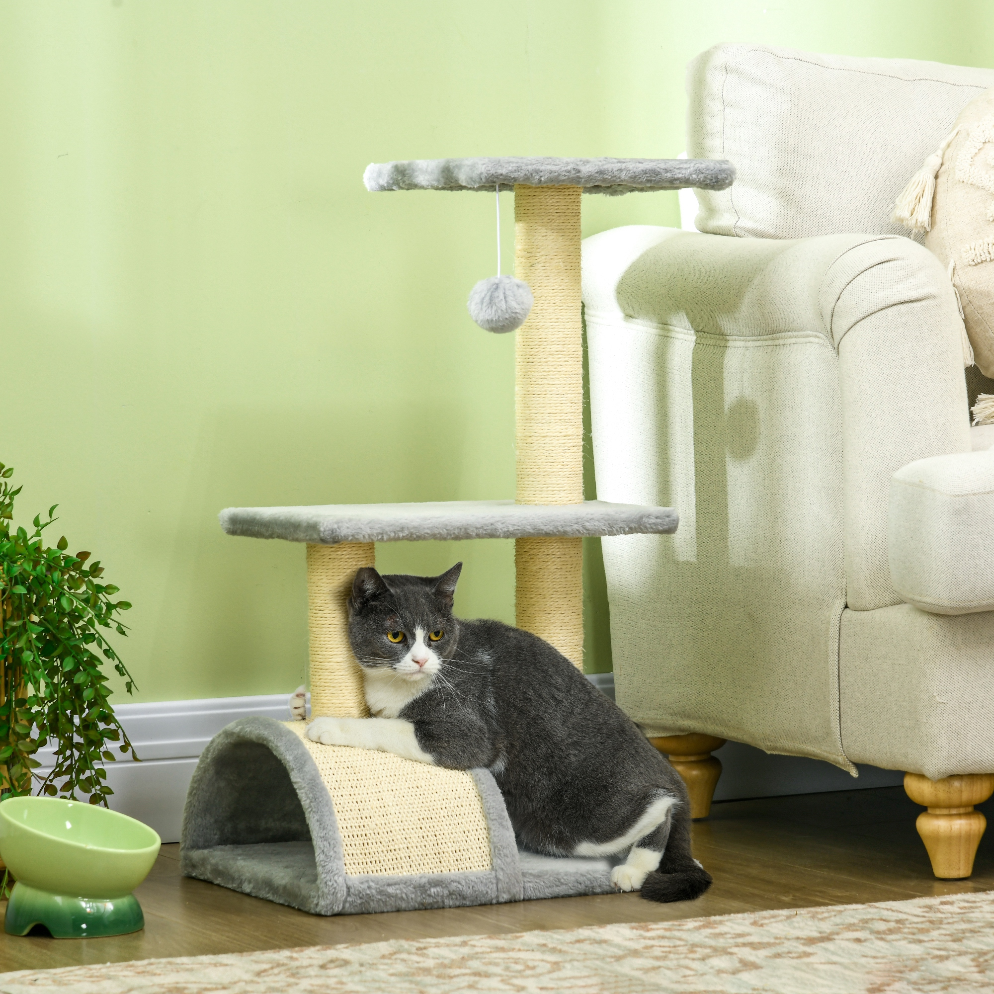 Un gatto grigio e bianco graffia un piccolo tiragraffi a più livelli vicino a un divano.