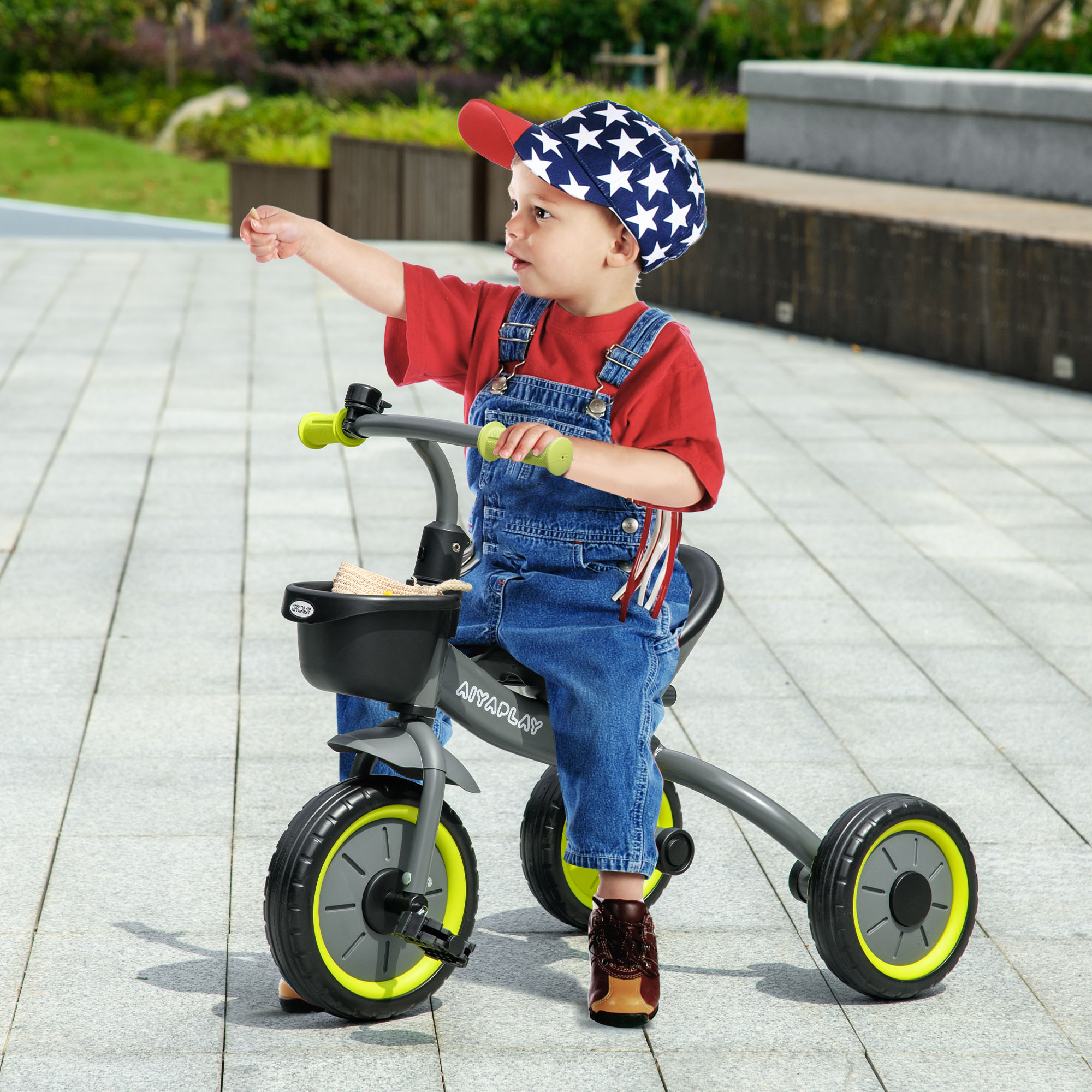 Un bambino con cappello stellato e salopette guida un triciclo su un vialetto pavimentato.