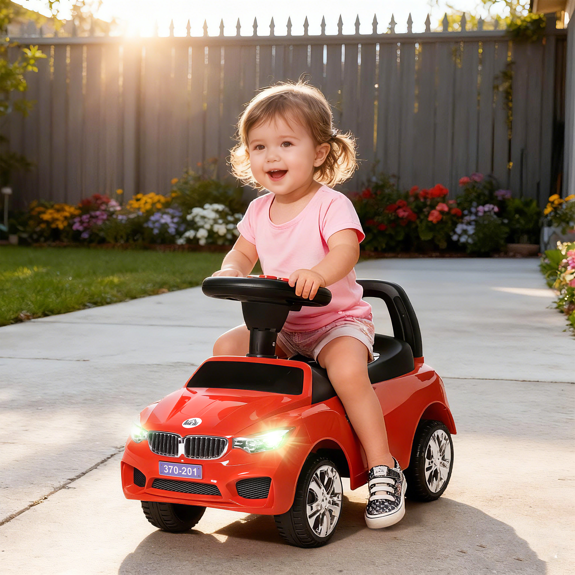 Bambino con camicia rosa sorride mentre guida una macchinina rossa su un vialetto soleggiato.