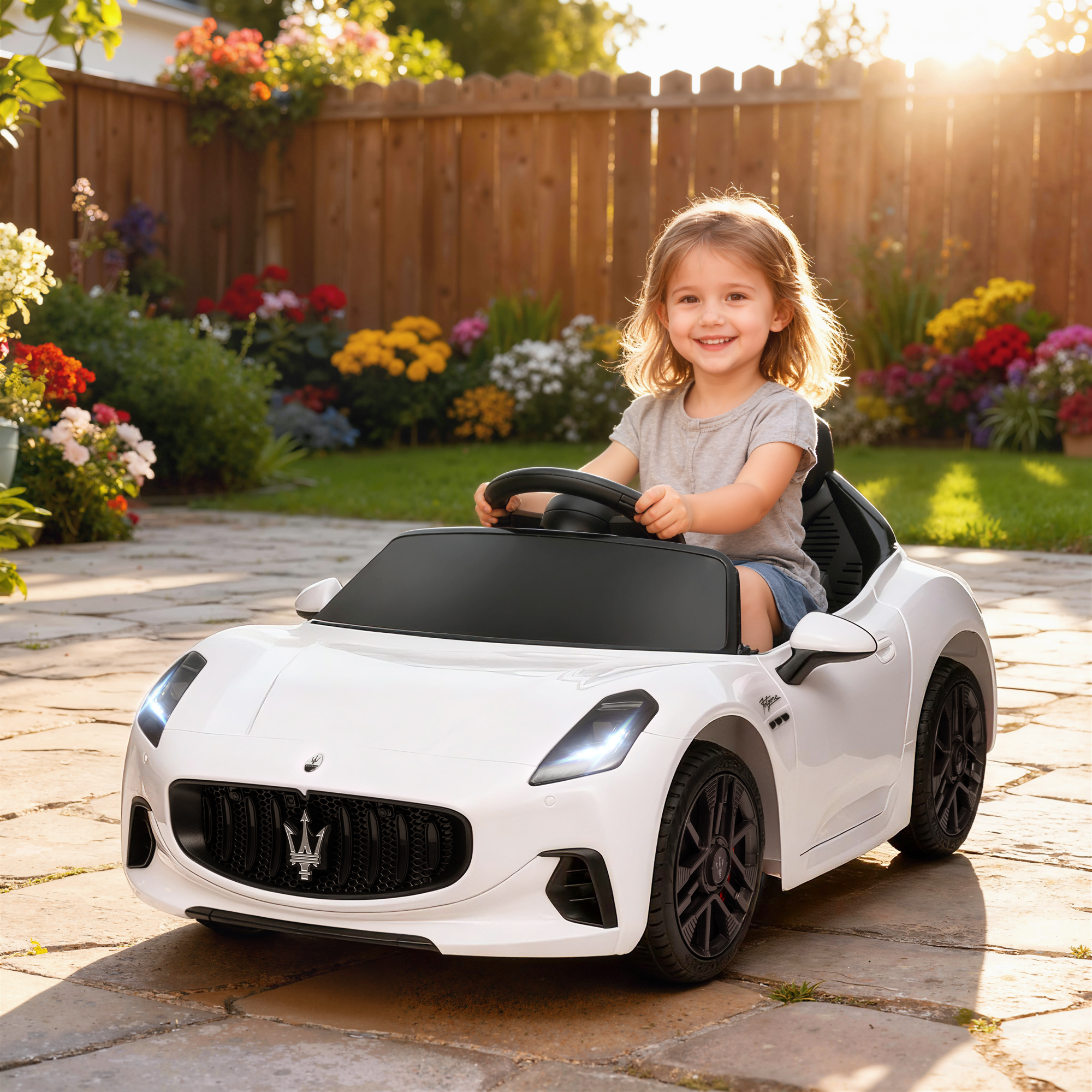Una bambina sorride seduta su un'auto giocattolo bianca in un patio soleggiato con fiori colorati sullo sfondo.
