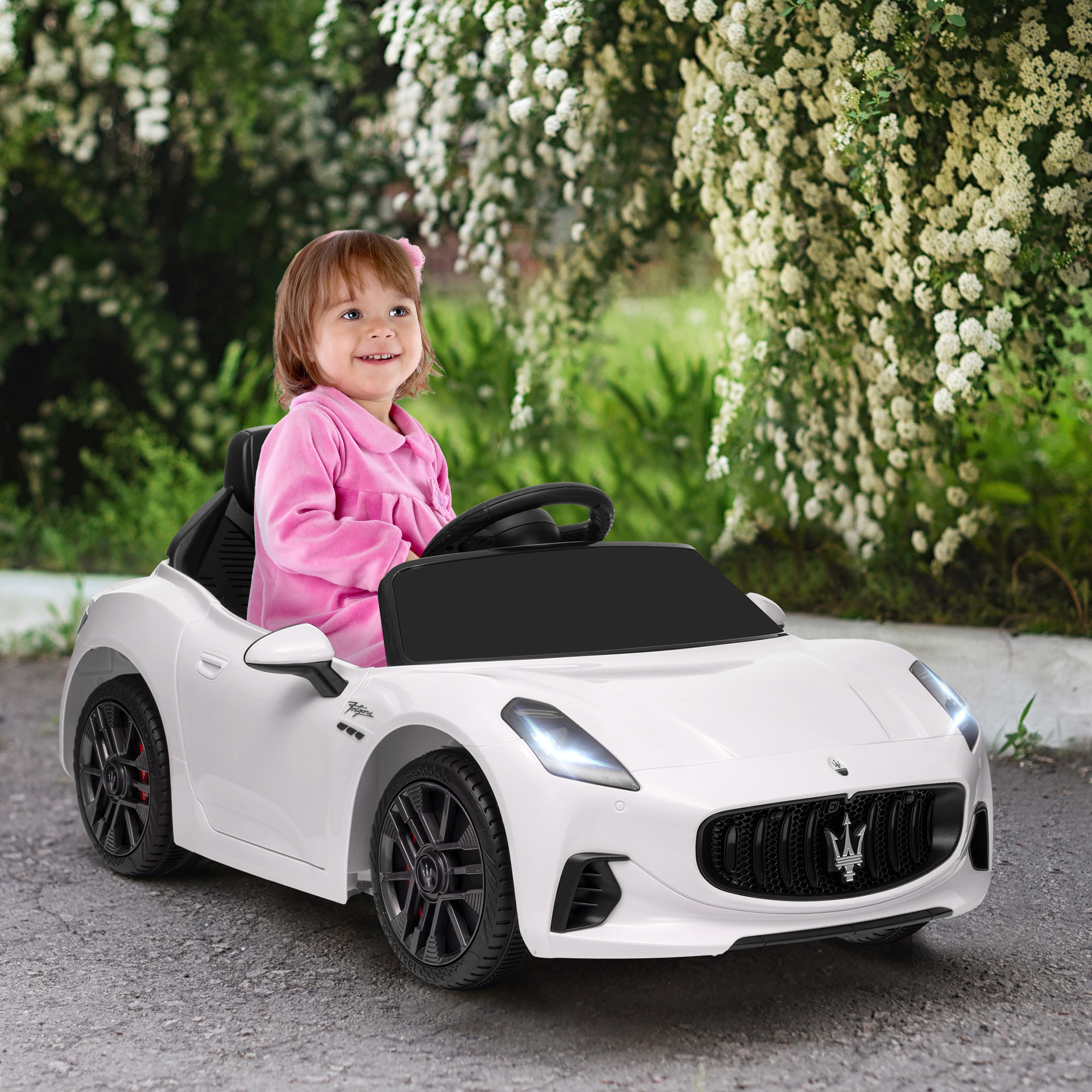 Una bambina sorridente in rosa siede su un'auto giocattolo bianca davanti a cespugli fioriti.