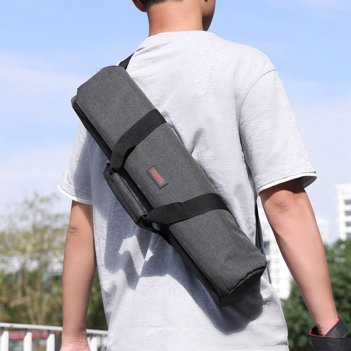 A person wearing a grey t-shirt carries a dark grey cylindrical shoulder bag outdoors.