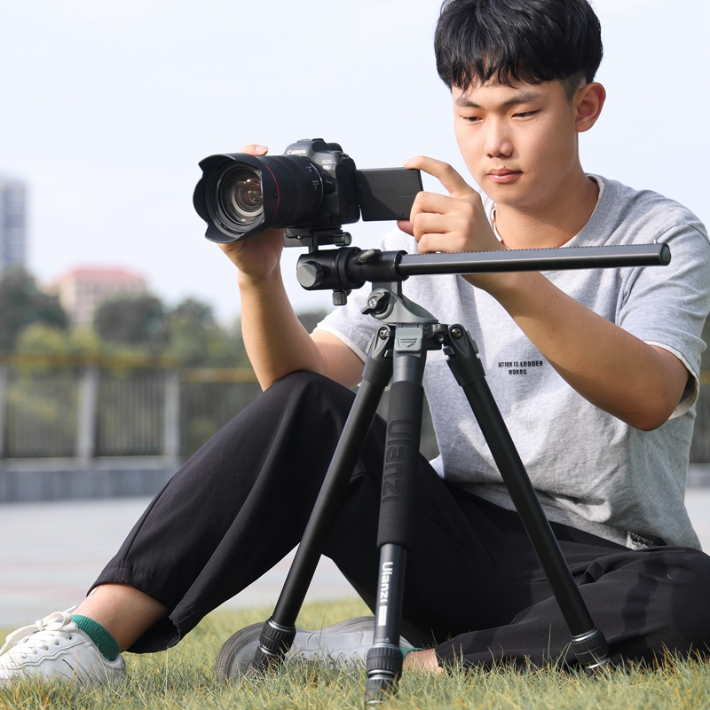 A young man sits on grass outdoors, adjusting a camera mounted on a black tripod.