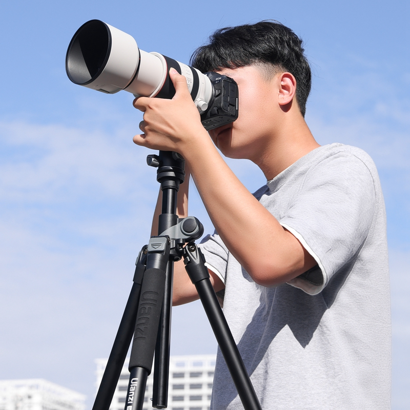 A young man in a grey shirt looks through a professional camera on a black tripod against a blue sky.